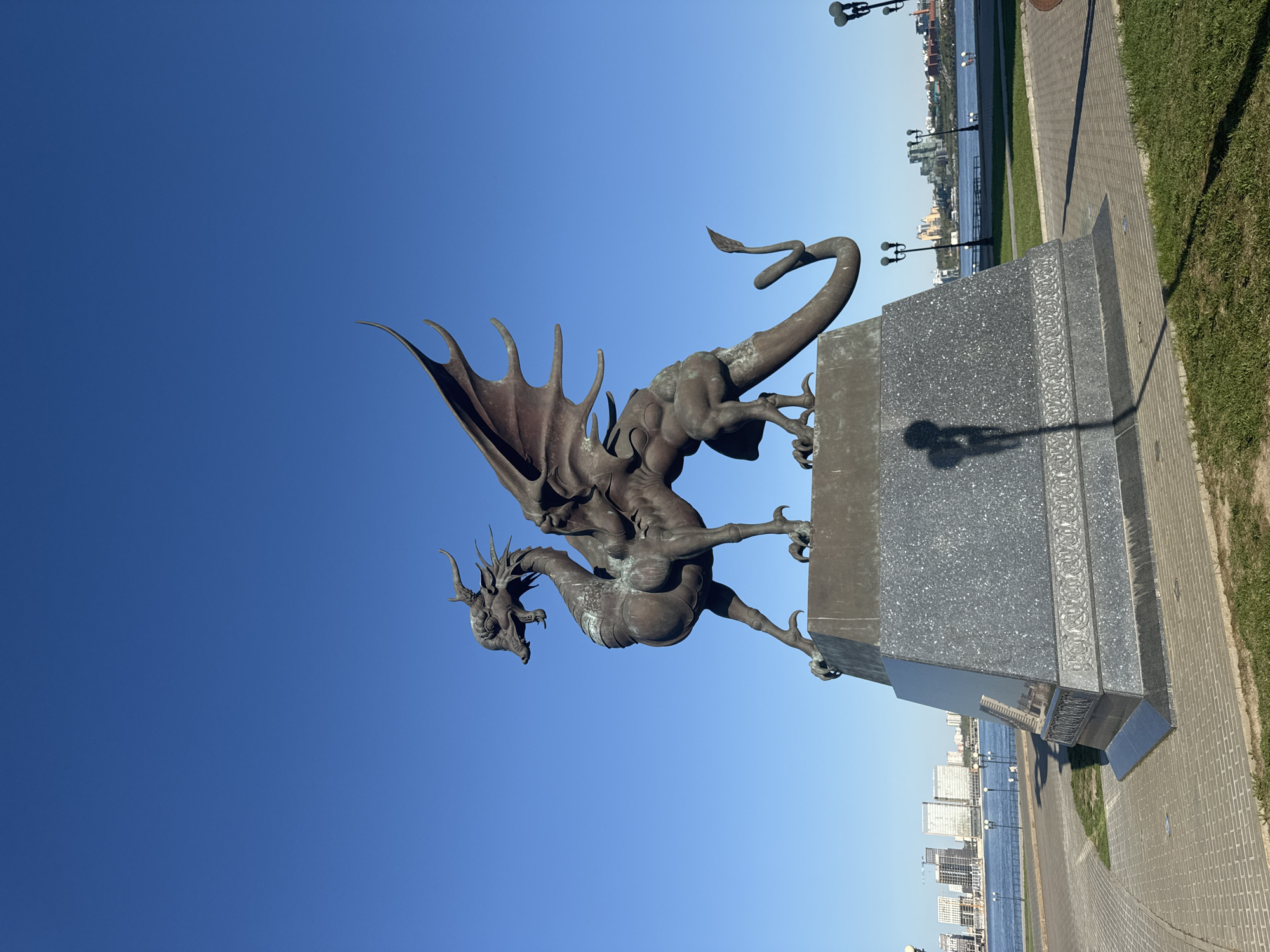 A full shot of the Zmey Gorynych dragon statue in Kazan, Russia. The dragon is made of metal and is perched on a granite base. The dragon is facing left, and its wings are spread. In the background, there is a clear blue sky and a river with buildings on the opposite shore. The statue is located on a brick sidewalk next to a grassy area. A street lamp is partially visible on the right.