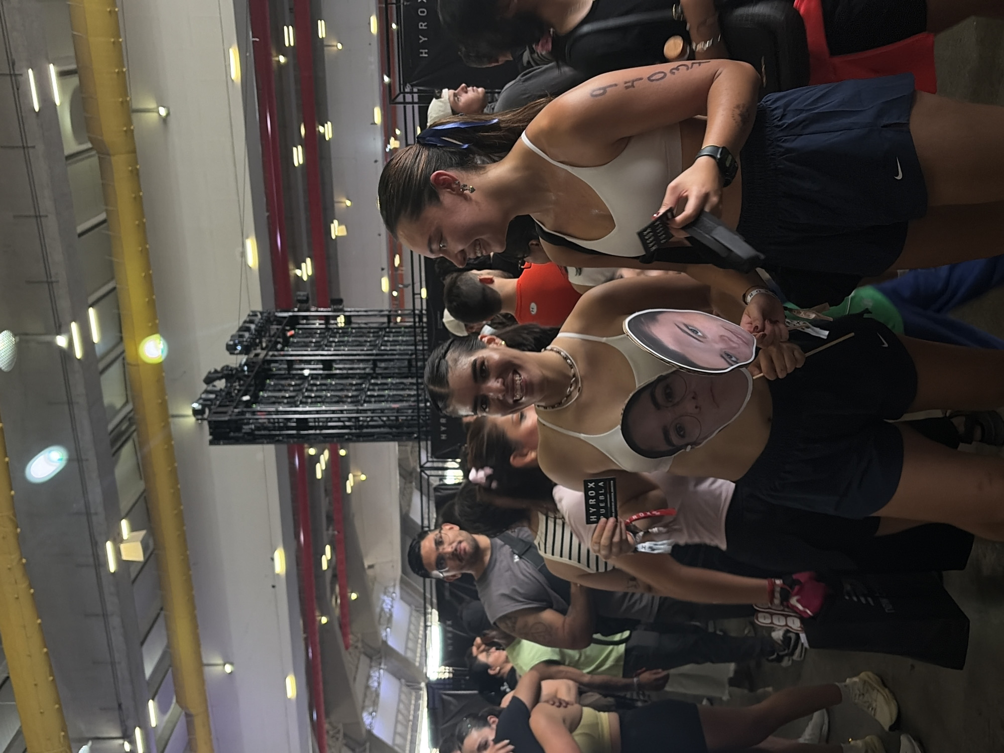 The image shows a group of people in an indoor space. Two women in the foreground are wearing athletic attire, including sports bras and shorts, with race numbers on their arms. One holds a cut-out face on a stick. Behind them, others are gathered, some with HYROX-branded materials. The environment features industrial-style ceilings with lighting fixtures, structural supports, and signage indicating the location as HYROX Puebla in the city of Unknown, Unknown.