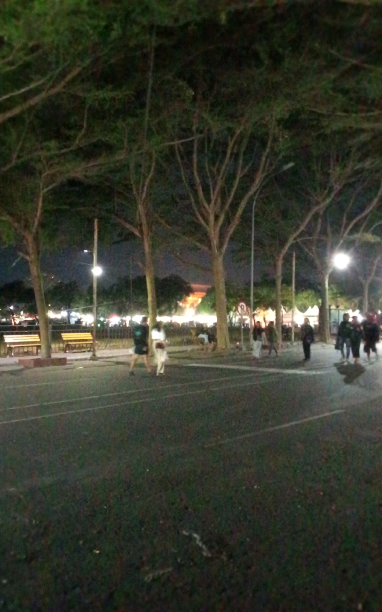 The image depicts a nighttime scene in the city of Ngasem, Indonesia, likely an outdoor public area such as a park or plaza, given the presence of trees, benches, and streetlights. In the foreground, a paved area, possibly a road or walkway, is visible with white lines. Several individuals are scattered throughout the scene. Two people are walking towards the right, one in a dark shirt and shorts, the other in a light-colored dress or skirt and top. Further to the right, a group of people are standing and walking, some appearing to be a family or friends. In the background, there are more trees and what appears to be a building with illuminated windows, suggesting it could be a commercial or public establishment. The overall atmosphere is dimly lit, illuminated by artificial lights from street lamps and the distant building.