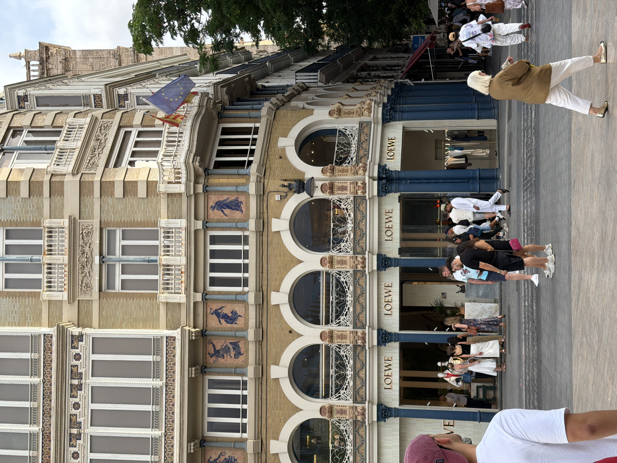 The image shows a street view of a building with a Loewe store on the ground floor. The building's architecture features multiple stories with ornate balconies, arched windows, and decorative tiling. Several people are visible on the sidewalk in front of the store, some browsing merchandise, others walking by. The sky is overcast. The location is stated as Unknown, Unknown.