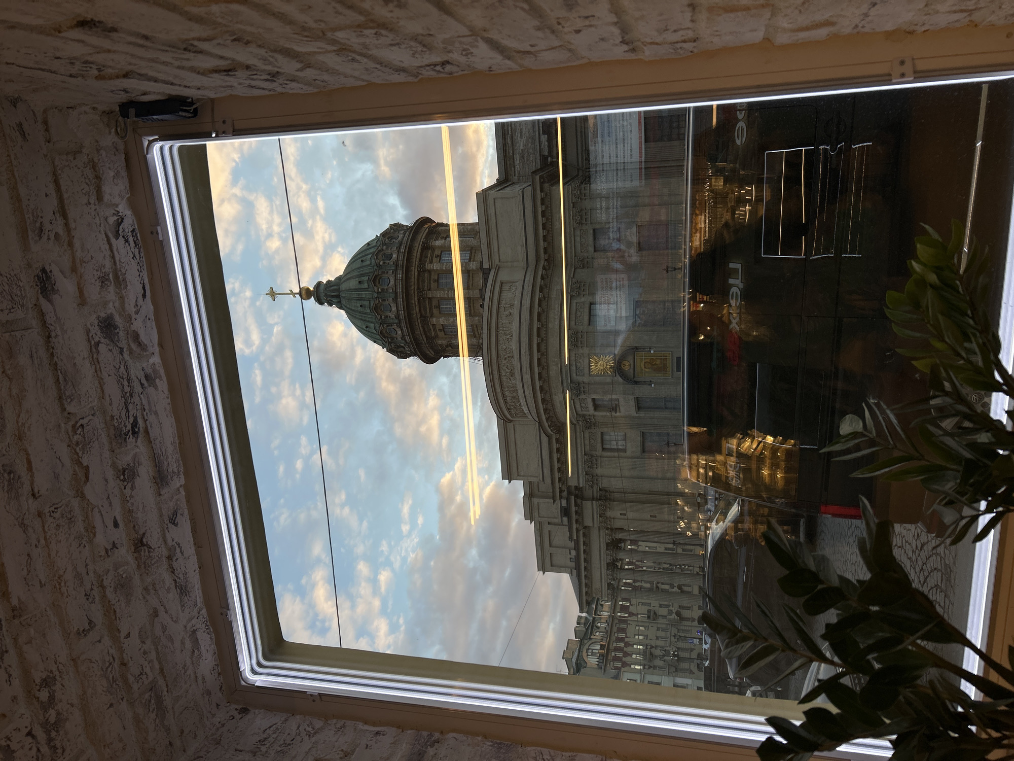 The image, taken from inside a building looking out a window, is oriented such that the viewer is looking up at the facade and dome of Kazan Cathedral in Saint Petersburg, Russia. The window frame, predominantly white, is visible along the edges, with a textured brick wall on the left and top, indicating an interior setting.
The main subject is the iconic Kazan Cathedral, with its large, green-domed roof culminating in a golden cross, prominently centered in the frame. The neoclassical architectural details of the cathedral's facade, including columns and intricate carvings, are also visible.
The sky outside is partly cloudy, displaying patches of blue sky interspersed with white and light orange clouds, suggesting either late afternoon or early evening light, possibly approaching sunset or twilight.
The lower right portion of the window is highly reflective. These reflections show street-level activity and structures from across the street. Visible in the reflections are parts of other buildings, the glow of artificial lights (likely streetlights or vehicle lights), and what appears to be the interior of the space from which the photo was taken, including a green, leafy houseplant in the bottom right corner.
Notable visible text, appearing in reflection on the window, includes Cyrillic letters that spell out parts of "ПЕРЕКРЕСТОК" (Perekrestok), indicating the presence of a store or advertisement for this Russian supermarket chain in the vicinity.
No specific people or direct human activity are depicted; the scene primarily captures architectural beauty and urban reflections during a tranquil time of day.