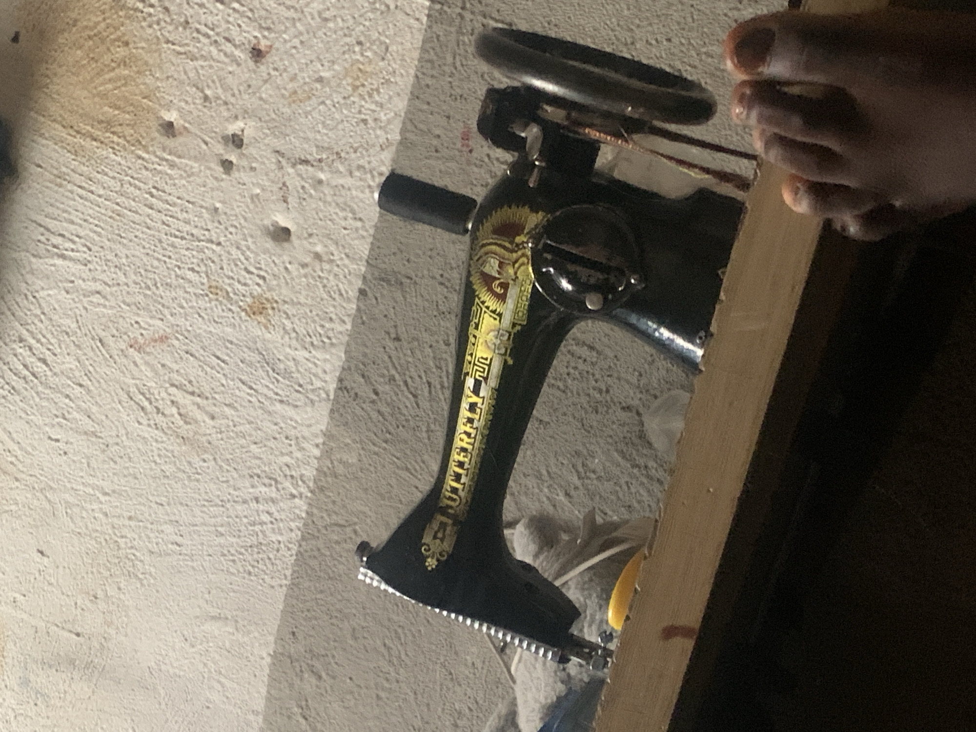The image depicts a vintage "Butterfly" brand sewing machine positioned on a wooden surface. The sewing machine is black with yellow and red detailing. The wall behind the sewing machine appears textured and is a light color with some brown stains and small holes. A person's foot, with orange-painted toenails, is visible in the lower right corner of the frame. The overall lighting suggests an indoor setting. The photograph was taken in Jauro Yinu, Nigeria.