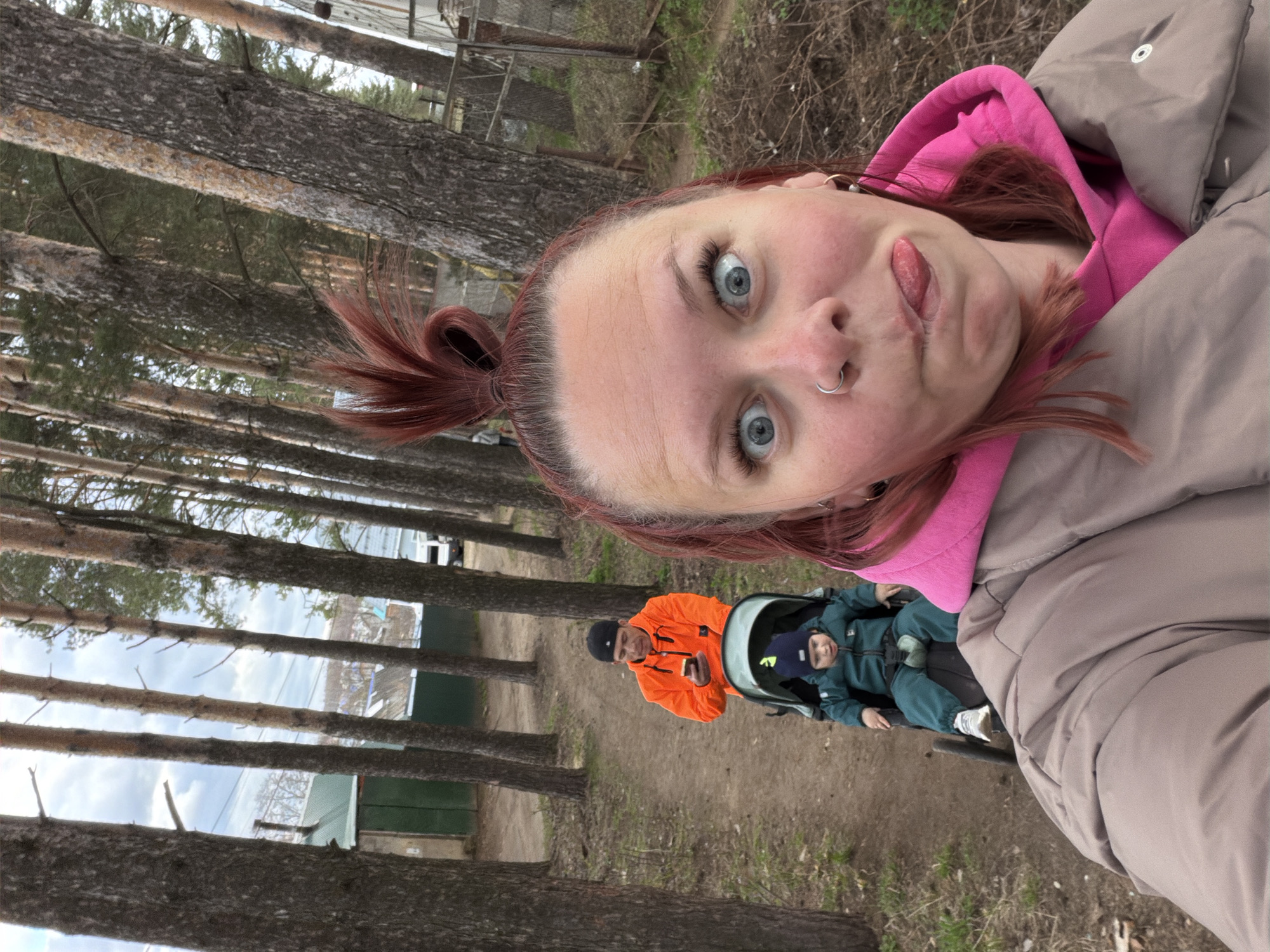 This selfie image captures an outdoor scene, likely in a park or wooded area in Chaikovsky, Russia, under an overcast sky.
In the foreground, a woman with reddish-brown hair tied in a high ponytail is taking the photo. She has bright blue eyes, a nose piercing, and is making a playful expression by sticking out her tongue. She is wearing a vivid pink hooded sweatshirt layered under a light beige or taupe puffer jacket.
Behind her, in the mid-ground, a man in a bright orange jacket and a dark beanie stands, looking towards the camera. He is holding a small, light-colored object, possibly a phone or food. Beside him, a young child is seated in a black and green stroller, dressed in a dark green or teal warm suit and a dark blue beanie.
The background features numerous tall, slender trees with rough, dark bark, characteristic of a pine forest. The ground is a dirt path with some patches of grass. Distant structures and glimpses of a cloudy sky are visible through the trees. The overall atmosphere suggests a casual outdoor outing on a cool, possibly chilly day. There is no visible text in the image.
