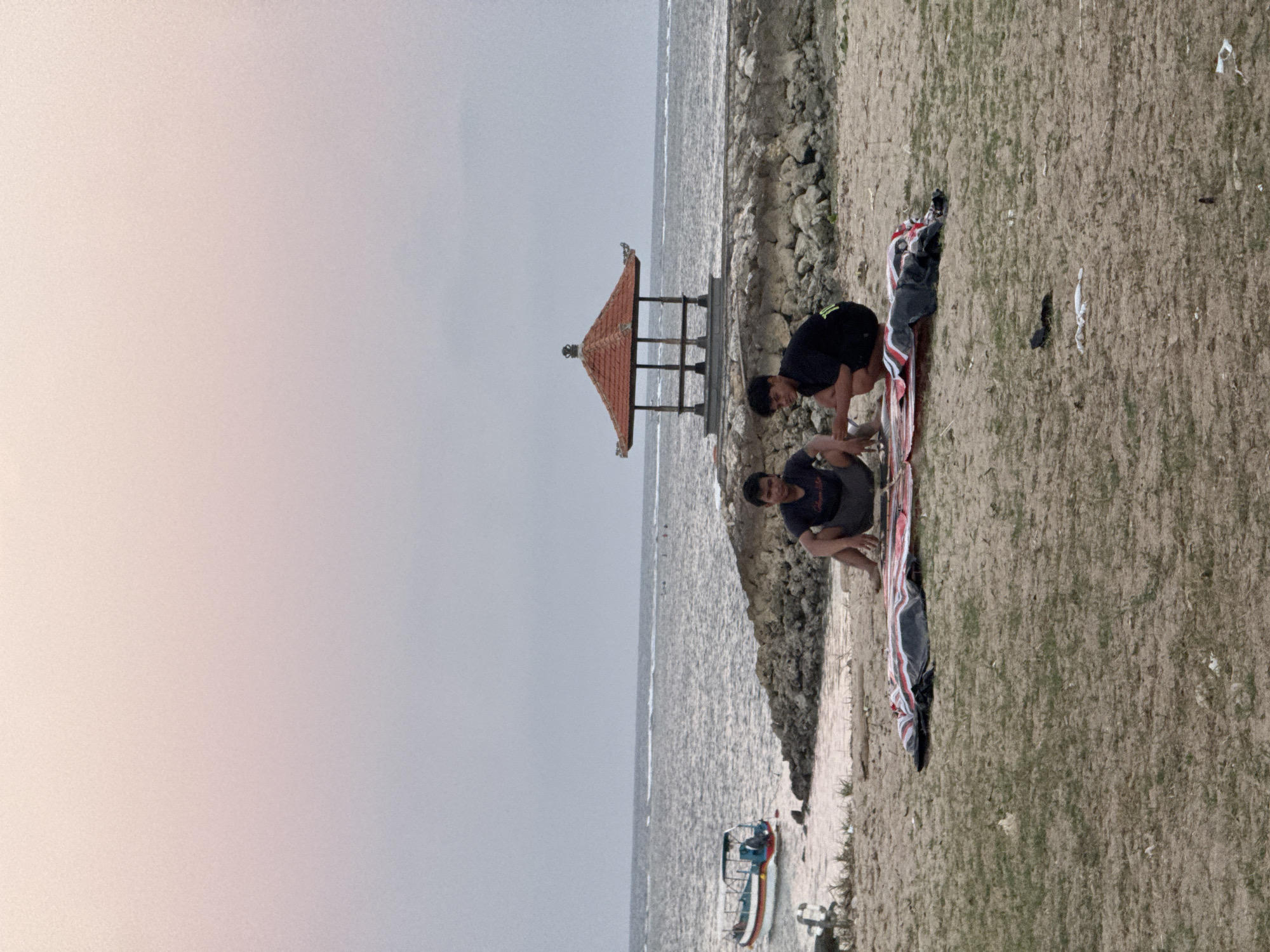 The image depicts a scene on a sandy beach with the ocean in the background. Two young men are the primary subjects, engaged in an activity on the sand. One man, wearing a black t-shirt and dark shorts, is in a crouched position, appearing to interact with the other man. The second man, also in dark attire, is positioned on a colorful mat or blanket, with his body partially visible. They are on a sandy area that transitions to rocky terrain towards the water.
In the middle ground, a traditional Balinese structure with a distinctive orange-tiled roof and wooden supports stands near the water's edge. The ocean is calm, with gentle waves reaching the shore. A small boat, painted with red and blue stripes, is docked near the beach.
The overall atmosphere suggests a relaxed daytime scene, possibly in the late afternoon given the soft lighting. The sky is overcast, indicating a cloudy or potentially misty weather condition. The location cues, such as the Balinese pavilion and the boat style, along with the provided location context, point to a coastal area in Kuta Selatan, Indonesia. There is no visible text in the image. The men's postures suggest concentration and engagement in their activity.