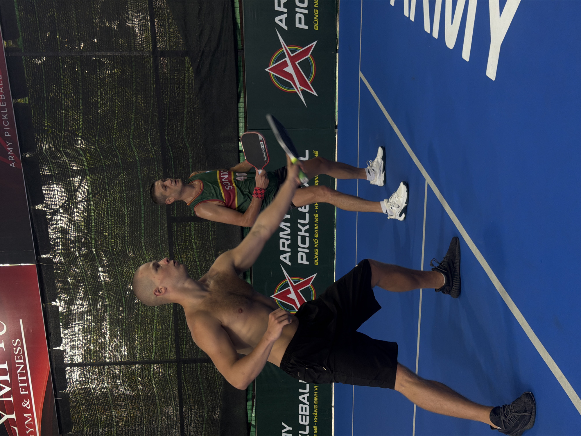Two men are playing pickleball on a court. One man is shirtless and wearing black shorts and black shoes. He is in the process of hitting the pickleball. The other man is wearing a green and red jersey with the word "SONIC" and the number "40" on it, white shorts, and white shoes. He is holding a pickleball paddle and watching the other man hit the ball. In the background, there is a fence, a banner that says "ARMY PICKLEBALL," and part of a sign that says "OLYMPIC GYM & FITNESS." The location is Bac Nha Trang Ward, Vietnam, as indicated by the Vietnamese writing on the banner: "BÙNG NỔ ĐAM MÊ" and "M MÊ - KHẲNG ĐỊNH VỊ."