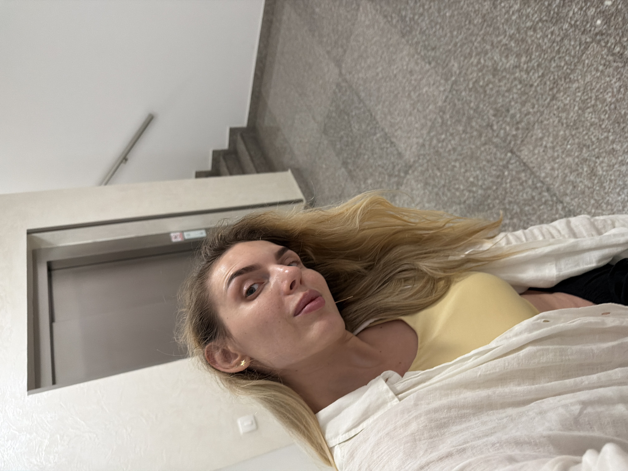 A blonde woman with light skin and full lips is taking a selfie indoors, leaning forward and looking towards the camera. She has on a yellow top and a white unbuttoned shirt. Her left ear, adorned with a star-shaped earring, is visible. In the background, there is a grey elevator door, a white wall, a metal handrail, and concrete stairs. The floor is made of grey granite tiles.