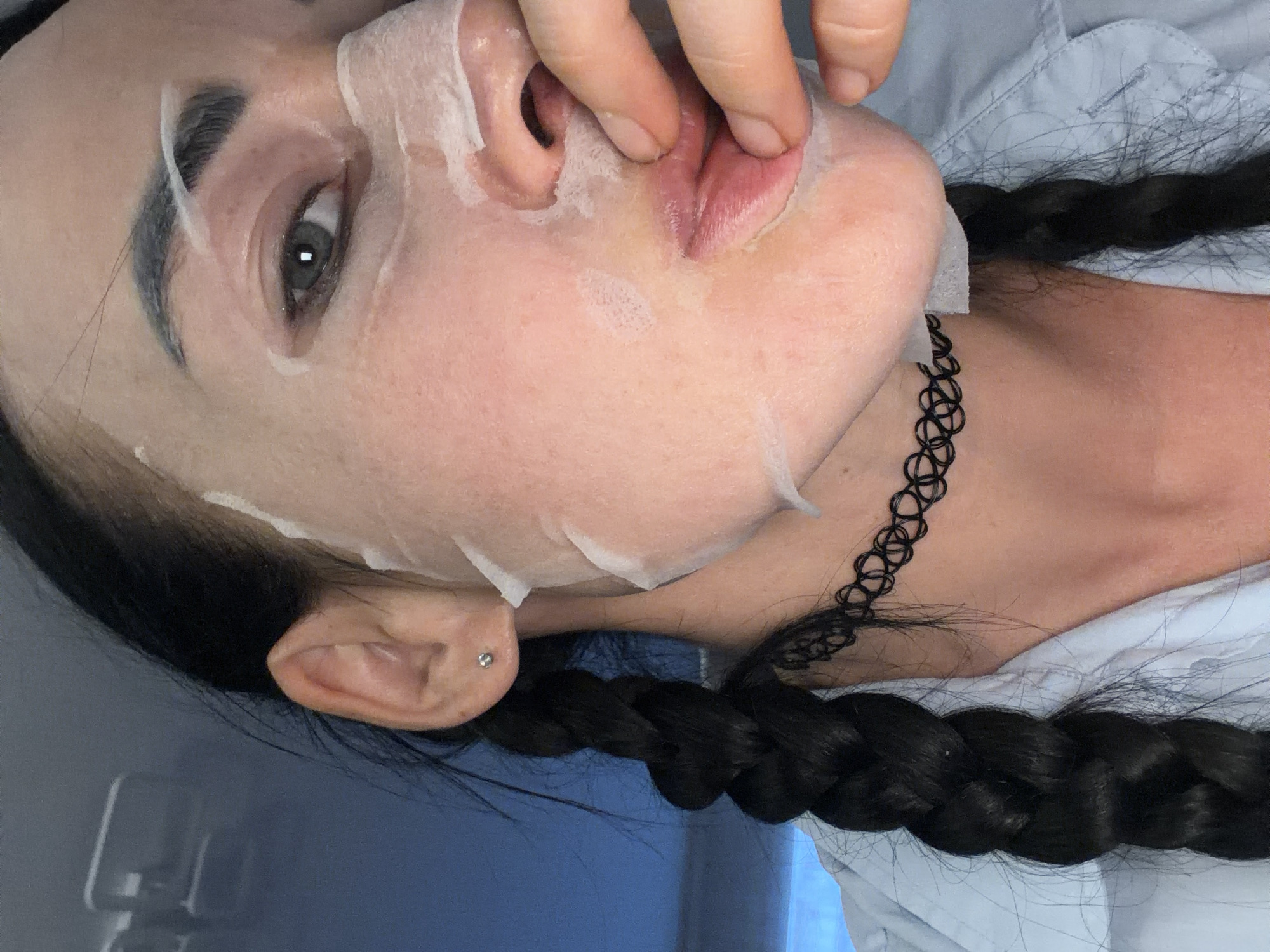 A close-up, medium-shot photograph shows a young Caucasian woman with dark braided hair and fair skin applying a sheet mask to her face. She is wearing a light blue collared shirt and a black choker necklace. The woman's left eye is visible, with dark eyebrows and dark eyelashes. Her right hand is touching her lips, which are covered by the sheet mask. The background is blurred and appears to be an indoor environment with a blue wall and some reflective surfaces. The image is taken from a slightly high angle.