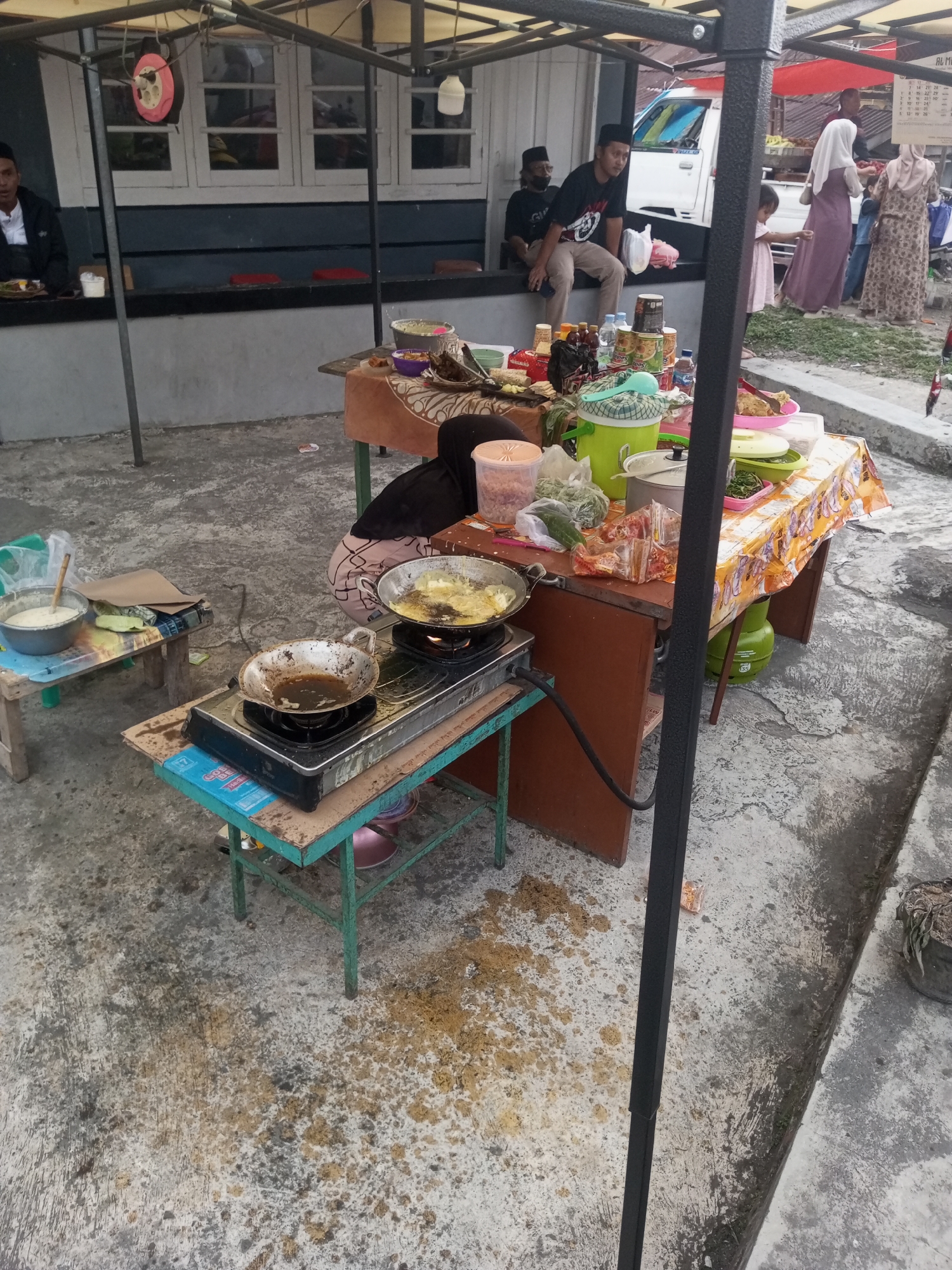 A street vendor is preparing food on a portable stove in the city of Pandansari, Indonesia. The vendor, wearing a black headscarf and apron, is standing behind a table laden with food items and cooking supplies. A frying pan on the stove contains what appears to be food being fried. The scene takes place outdoors, with a concrete ground surface and a canopy providing shade. In the background, other people are visible, some sitting and some standing. A white truck and various stalls suggest a marketplace or a bustling roadside area. The overall impression is of a lively outdoor food stall environment.