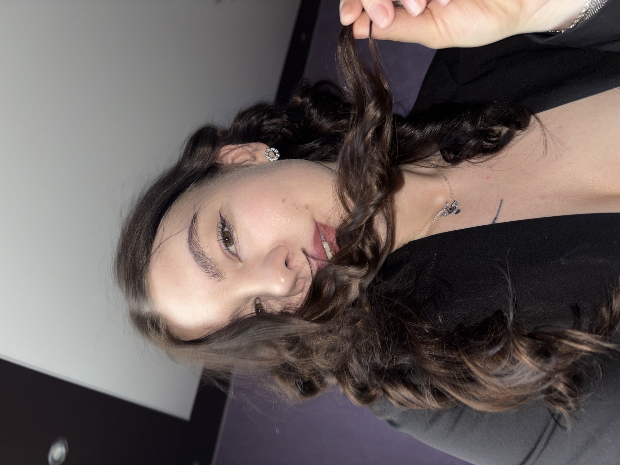 A woman with brown curly hair is positioned at an angle, her head closer to the top of the frame. She wears a black top. The background is a mix of grey and black, likely a wall. She has fair skin and visible jewelry, including earrings and a necklace. Her hand is visible at the top, holding a strand of her hair. The city of Perm, Russia, is not discernible from the image.