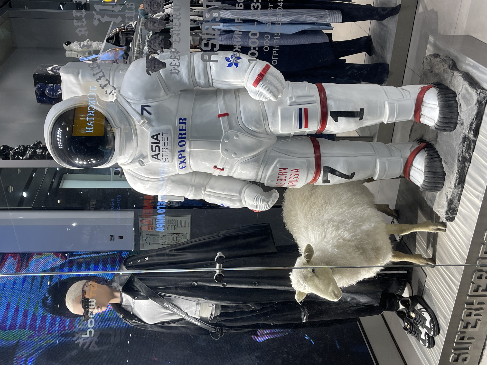 The image shows a store window display featuring an astronaut mannequin lying on its side, dressed in a white spacesuit with the words "ASIA STREET EXPLORER" and "AS BORN RUSSIA" printed on it. There are also numbers "71", "1", and "7" visible on the suit. Next to the astronaut is a taxidermied sheep. Below these figures is another mannequin dressed in street style clothing, including a black leather jacket. The display is likely located in a shopping center, possibly in Saint Petersburg, Russia, judging from the Cyrillic text and other signs in the background. The store's logo appears to be "SUPERHEROES". The scene also shows a reflection of the mall environment, including lighting and other store displays.