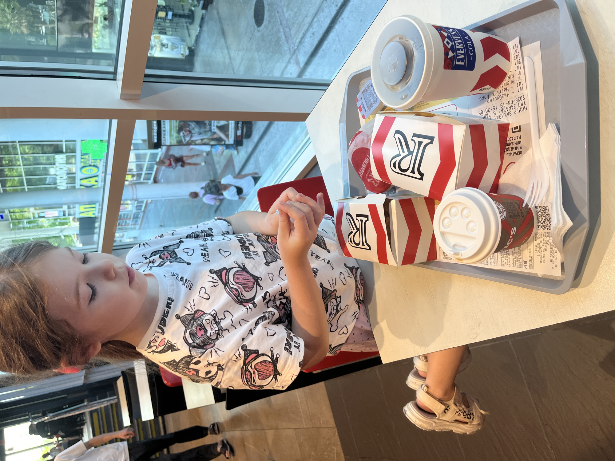 In a fast-food restaurant in Sochi, Russia, a young girl with blonde hair is sitting at a table by a large window, looking down. She is wearing a white t-shirt with a cartoon print. In front of her, on a grey tray, is a meal consisting of two red and white paper bags, likely containing food items, and a white paper cup with a red and white logo that reads "EVERVESS COLA". There is also a receipt and a plastic fork and knife. Through the window, the bustling street scene of Sochi can be seen, with people walking by and businesses with signs, including one that says "МАТОЛОГИЯ" (Dentistry). The lighting suggests it is daytime, possibly late afternoon, given the strong shadows. The overall scene depicts a casual meal break. The receipt contains Russian text and dates such as "2026-06-19", indicating the time of purchase.