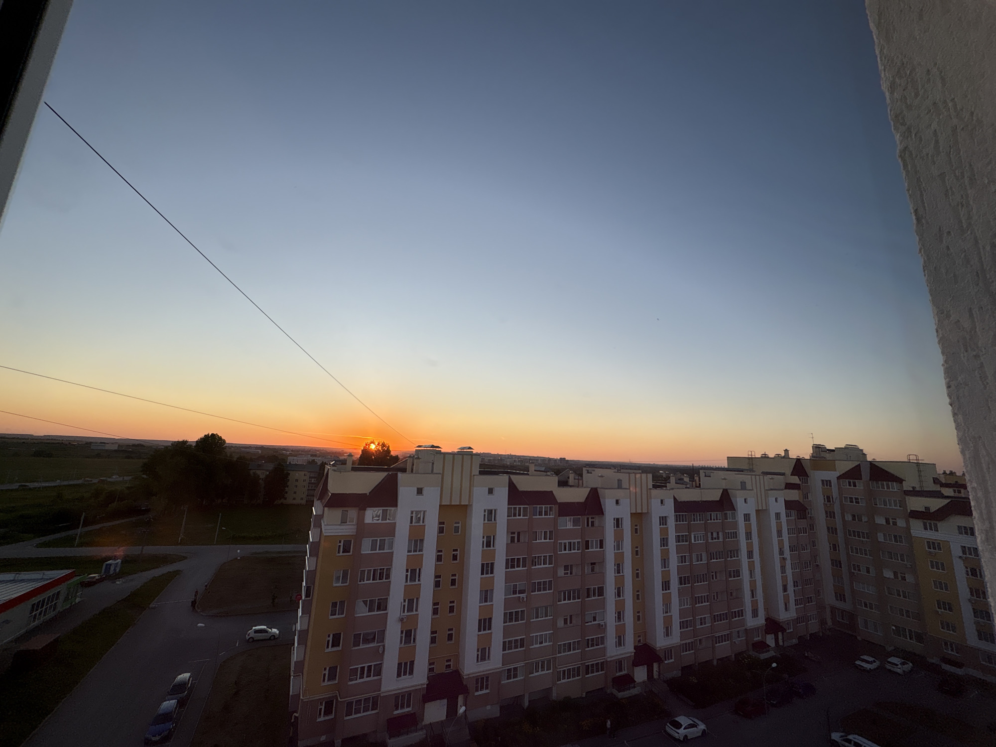The image depicts a panoramic view of a residential area at sunset, likely in the city of Засечное, Russia, given the context provided. The sky transitions from a pale blue at the top to warm oranges and yellows near the horizon, indicating the sun is setting. A single, thin, dark line, possibly a power line or wire, stretches diagonally across the upper portion of the sky.
In the foreground, on the right side, a rough, textured white wall occupies a significant portion of the frame. To the left, a long, multi-story apartment building dominates the mid-ground. This building has a varied facade with sections in yellow, beige, and reddish-brown, featuring numerous windows with white frames. The building is L-shaped or has multiple connected sections.
Beyond the apartment building, a street network is visible, with a few cars parked and driving. One white car is near the apartment building, and several other cars are parked on the street. A smaller building with a red and white facade, possibly a commercial establishment, is located to the left. The background consists of more buildings, trees, and a flat, open landscape stretching towards the horizon, all bathed in the soft light of the setting sun. The overall scene captures a quiet, urban evening.