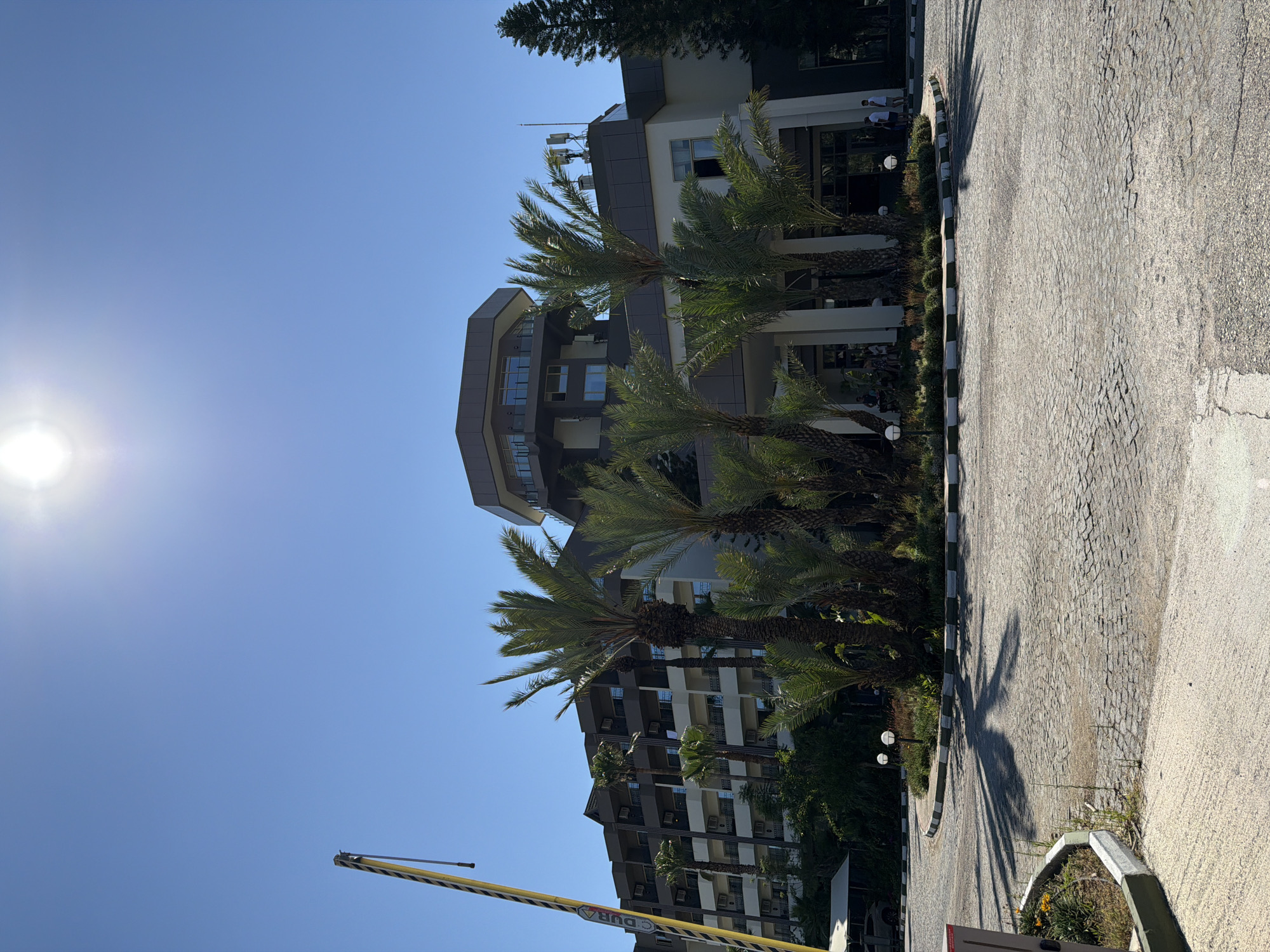 The image captures a sunny day scene in front of a modern, multi-story building. The building, with its sleek design featuring large windows and balconies, is partially obscured by several palm trees. The sky is a clear, bright blue, indicating fair weather. The sun is visible in the upper left corner of the image, casting long shadows from the palm trees onto a paved area that appears to be a driveway or walkway. The paving is made of cobblestones in some areas and a lighter, smoother material in others.
There are a few people visible in the background, near the entrance of the building, appearing to be walking or standing. There is also a yellow and black striped barrier or gate in the foreground, with some text on it. Although the text is partially obscured and blurry, "DURA" is legible. The overall impression is one of a warm, perhaps tropical or Mediterranean, location during daytime. The scene is peaceful and well-lit, suggesting a pleasant atmosphere.