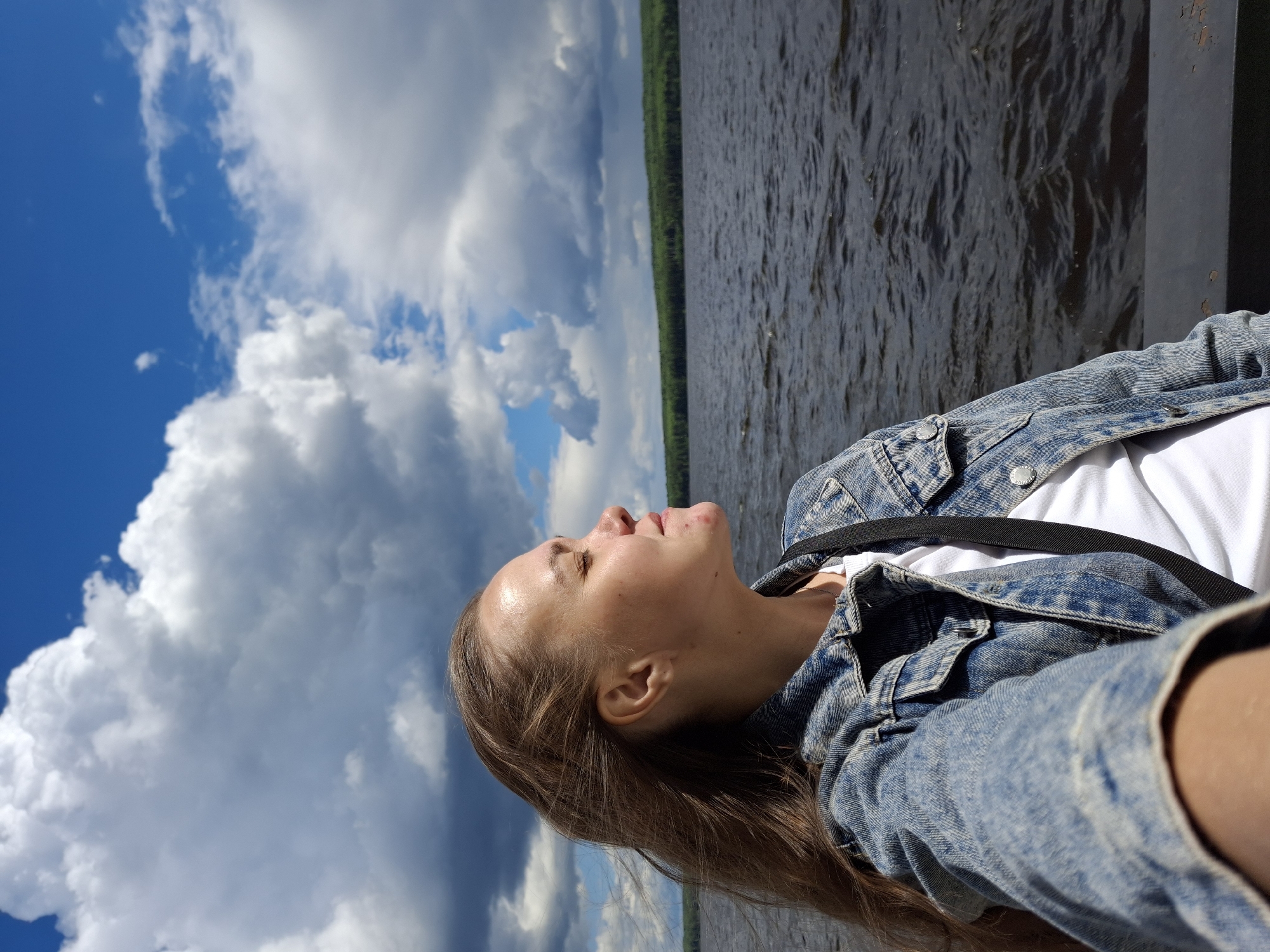 A photograph shows a woman looking up at the sky with puffy clouds. The background features a body of water, possibly the Kama River, with a forest line visible on the opposite shore. The woman is wearing a denim jacket over a white shirt. The image appears to have been taken near the city of Chaikovsky in Russia, given the local vegetation.