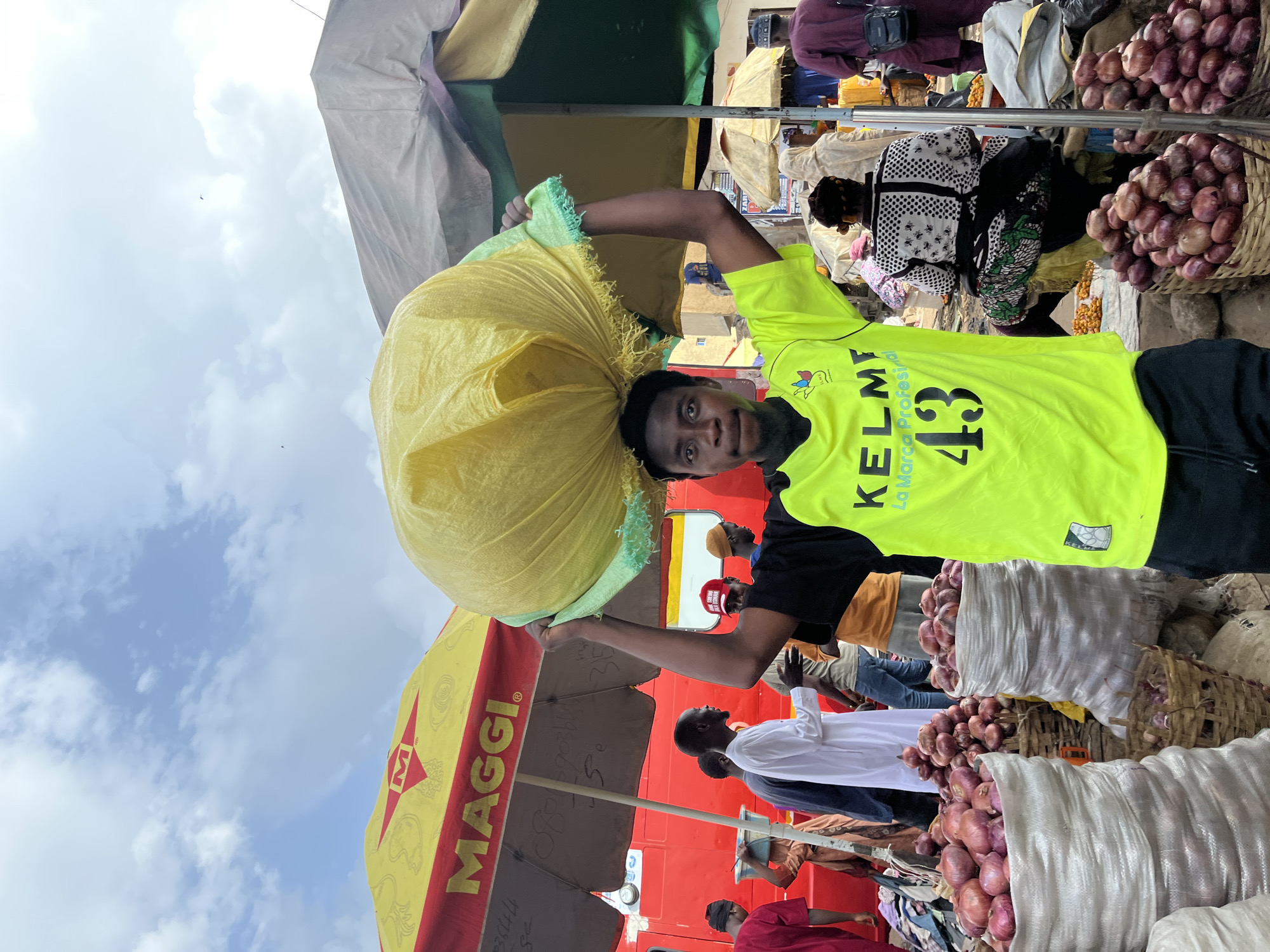 This media file captures a bustling market scene in Rafin Par, Nigeria. The primary subject is a young man carrying a large, yellow sack of goods on his head. He is wearing a bright neon green shirt with the word "KELME" and the number "13" visible, along with some smaller logos. Behind him, the market is filled with activity.
Several other people are present, some engaged in selling or buying, while others are simply part of the crowded environment. In the background, there are stalls and displays of produce, notably large baskets and sacks filled with red onions. A red vehicle, possibly a truck, is visible behind some of the market stalls, with a yellow awning featuring the "MAGGI" logo.
The setting appears to be an outdoor market, possibly under some form of shelter or canopy, given the presence of awnings and the overhead view of the sky with clouds. The time of day is likely daytime, indicated by the natural light and the activity. The overall atmosphere seems lively and busy, characteristic of a market. There is no clear indication of specific emotions, but the focus on the man carrying his load and the abundance of goods suggests a scene of commerce and daily life.