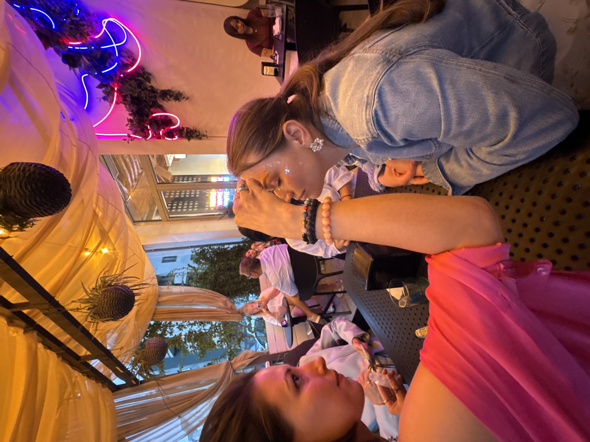 This image captures an intimate evening scene at a bar in Montenegro, featuring several patrons socializing under decorative lighting. The photo is taken from a low angle, looking up and across a table.
In the foreground, a woman with dark hair, wearing a bright pink top, looks towards the mid-ground. Her left arm, adorned with rings, is visible, holding a glass with ice. Next to her, another woman with brown hair, wearing a denim jacket, is leaning forward with her head bowed, her eyes looking down. She has glitter scattered on her forehead and cheekbones, and is wearing intricate pearl earrings and a stack of beaded bracelets on her left wrist.
In the background, other patrons are visible, including a woman in a maroon top seated at a table. The setting is adorned with various decorative elements: warm, draped fabric creates canopy-like sections, illuminated by string lights. Above, a vibrant pink and blue neon sign, integrated with some greenery, adds a modern, artistic touch. Large, spiky artificial flowers resembling proteas hang as part of the decor. Through windows or open spaces, glimpses of what appears to be trees and other buildings can be seen, suggesting an outdoor or semi-outdoor patio setting.
The ambiance is dim and cozy, characteristic of an evening establishment. On a reflective surface in the background, faint text is visible, including "YOU'RE COOL" and "YOU'RE WELCOME," likely part of a decorative element. The overall impression is one of a relaxed social gathering in a stylish, well-decorated bar.