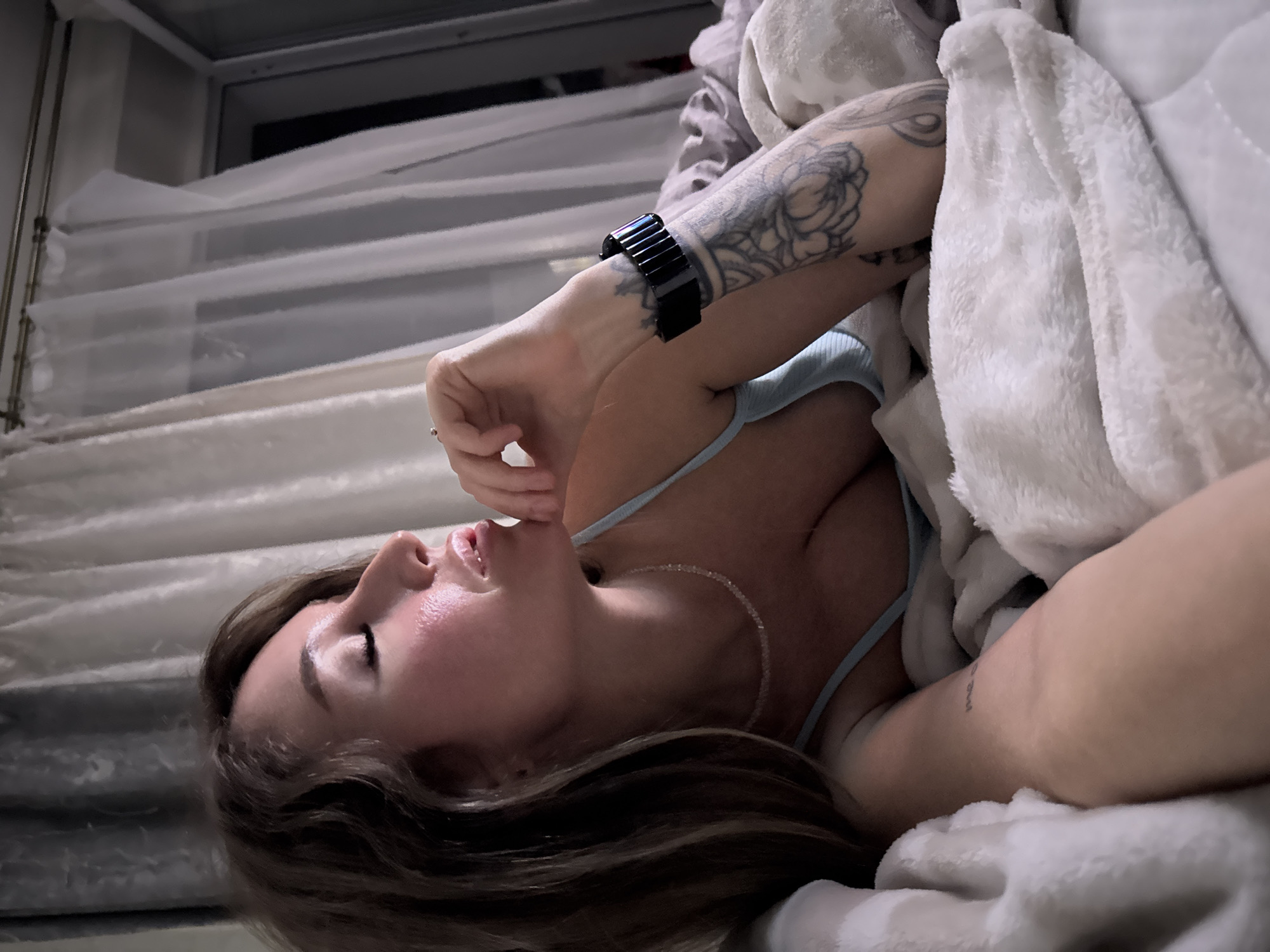 A woman with brown hair lies in bed, her eyes closed and her head resting on her hand. She is wearing a light blue camisole and a beaded necklace. A large tattoo is visible on her left arm, and she is wearing a black smartwatch. The bed is covered in white blankets, and sheer curtains are visible in the background. The scene appears to be indoors, possibly in a bedroom. The location is not specified in the image itself, and it is impossible to determine if it is in the city of Chaikovsky, Russia.