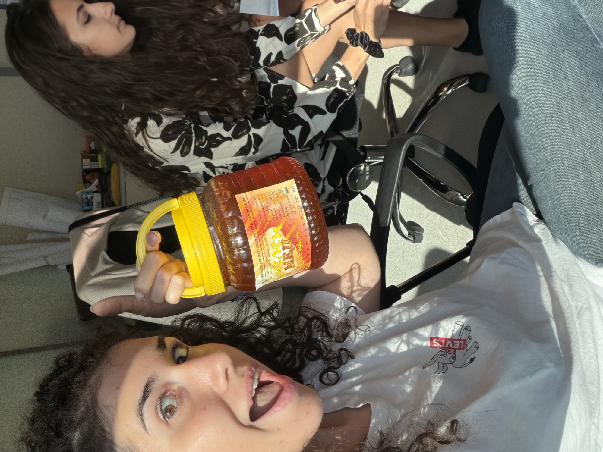 Two young women are indoors, possibly in an office or home setting, with sunlight streaming in from the right. The woman in the foreground, with curly dark hair and wide-eyed expression, is holding a large jar of honey with a yellow lid. She is wearing a white t-shirt with a red and black graphic of an animal and the word "LEVI'S". She is looking towards the camera with her mouth open. The woman in the background, also with long dark curly hair, is wearing a black and white floral print shirt. She is looking downwards and to her right, and her hands are partially visible. The environment appears to be an indoor space with office chairs and what looks like shelves with various items in the background, suggesting a place in Sofia, Bulgaria. The lighting indicates it is daytime.