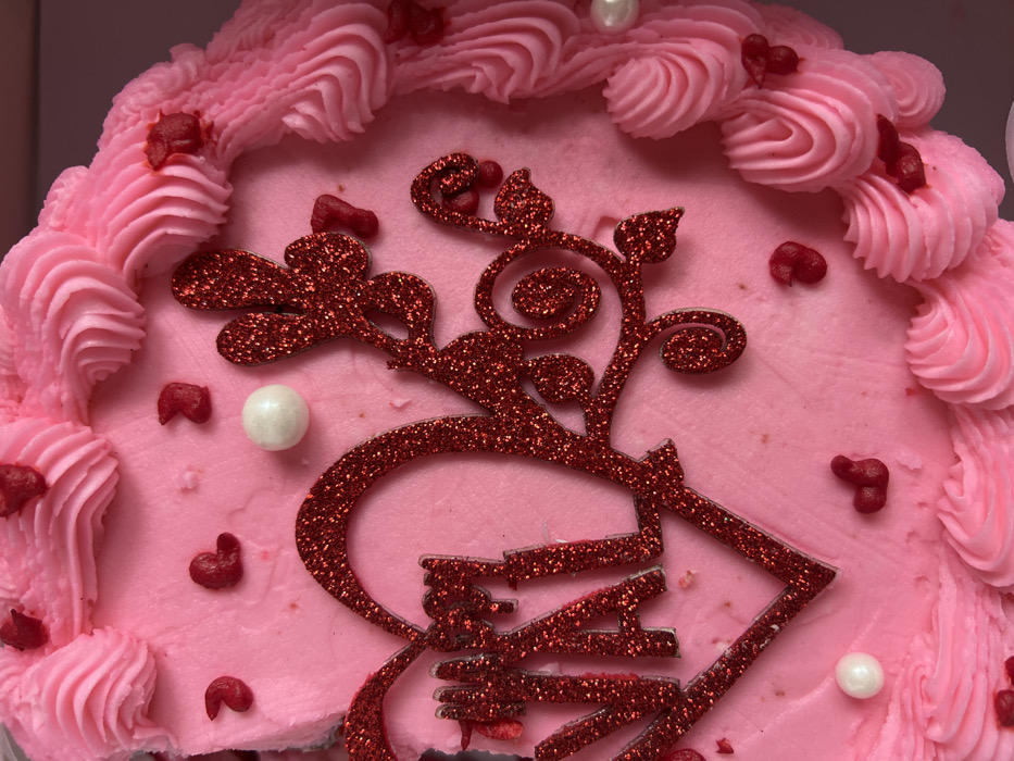The image displays a top-down view of a round cake, predominantly decorated in shades of pink and red, giving it a festive and sweet appearance.
The cake is covered in a light pink frosting. The top surface is smooth and evenly spread, while the perimeter features an ornate border of piped pink frosting. This border consists of numerous swirled rosettes and ridged S-curves, creating a volumetric, ruffled texture around the edge of the cake.
A large, intricate decoration, made of shimmering red glitter, is centrally placed on the smooth pink frosting. This glitter piece features stylized, scrolling branches and leaves, giving it an organic, vine-like quality. Integrated into the lower part of this glitter design, in a bold, decorative font, is the word "LOVE". The glitter material gives this main decoration a sparkling, textured look.
Scattered across the smooth pink frosting surface and nestled amongst the piped border are various smaller decorations. Many of these are small, irregularly shaped pieces of red candy or icing, some clearly resembling miniature hearts. There are also a few smooth, spherical white pearls, offering a contrasting color and texture to the red and pink elements.
The overall color palette of pink, red, and white, combined with the heart shapes and the word "LOVE", strongly suggests the cake is for a romantic occasion, such as Valentine's Day, or a celebration of love. The lighting appears bright and even, highlighting the details and textures of the frosting and decorations.