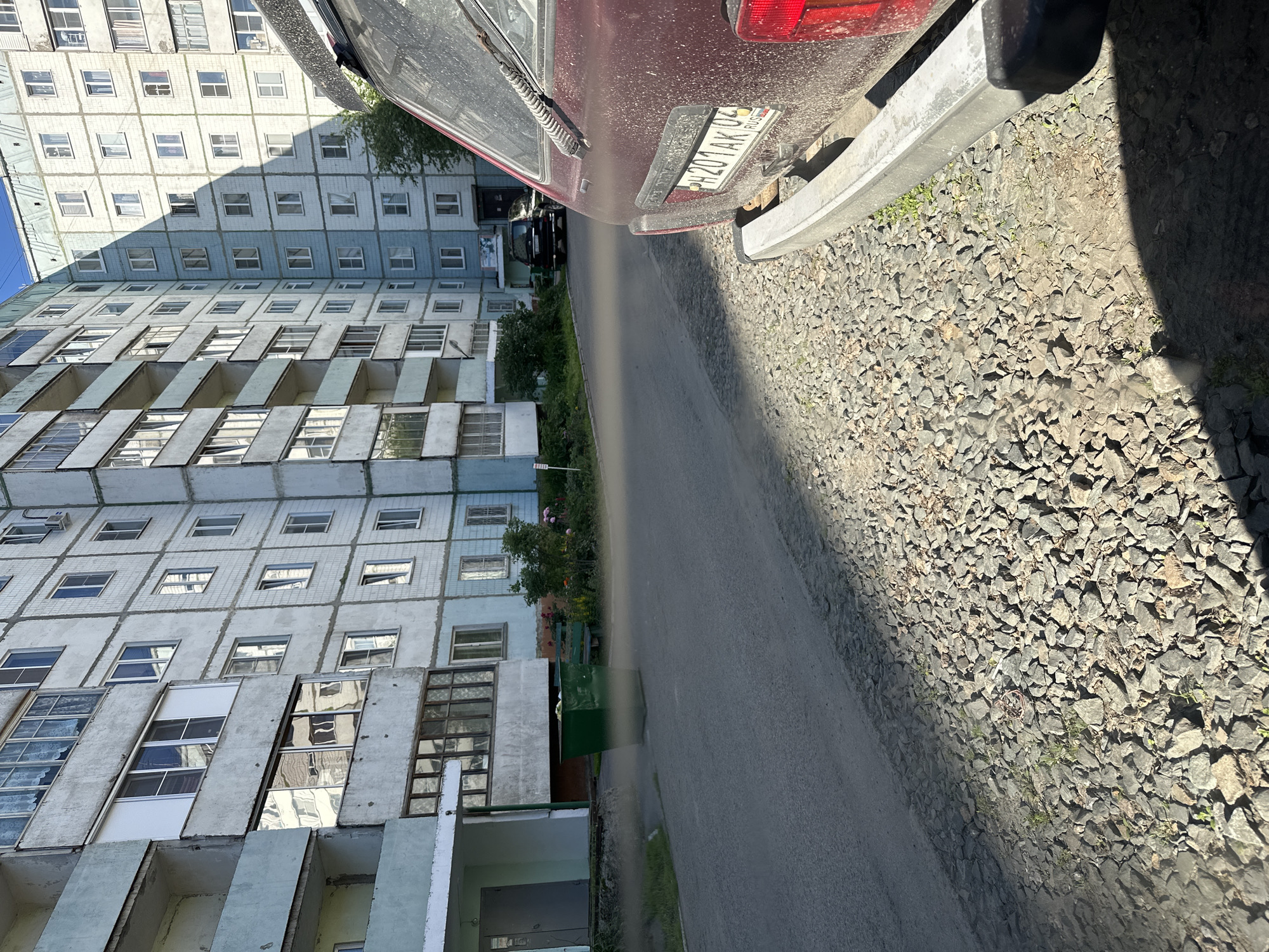 This image captures a street scene in Novoaltaysk, Russia, with a focus on residential buildings and parked vehicles. A multi-story apartment building, typical of Soviet-era architecture, dominates the background. Its exterior is light blue and gray, with numerous windows and balconies. In the foreground, the rear of a dusty, dark red vehicle is visible, with its license plate readable as H201AK04 RUS. The vehicle is parked on a gravelly shoulder next to an asphalt road. Further down the road, another dark-colored vehicle is parked in front of the apartment building, and a green trash bin is positioned near the entrance to one of the building's sections. A small green bench and some vegetation, including flowering plants and trees, are visible near the trash bin. The scene appears to be a typical urban environment with clear daylight.