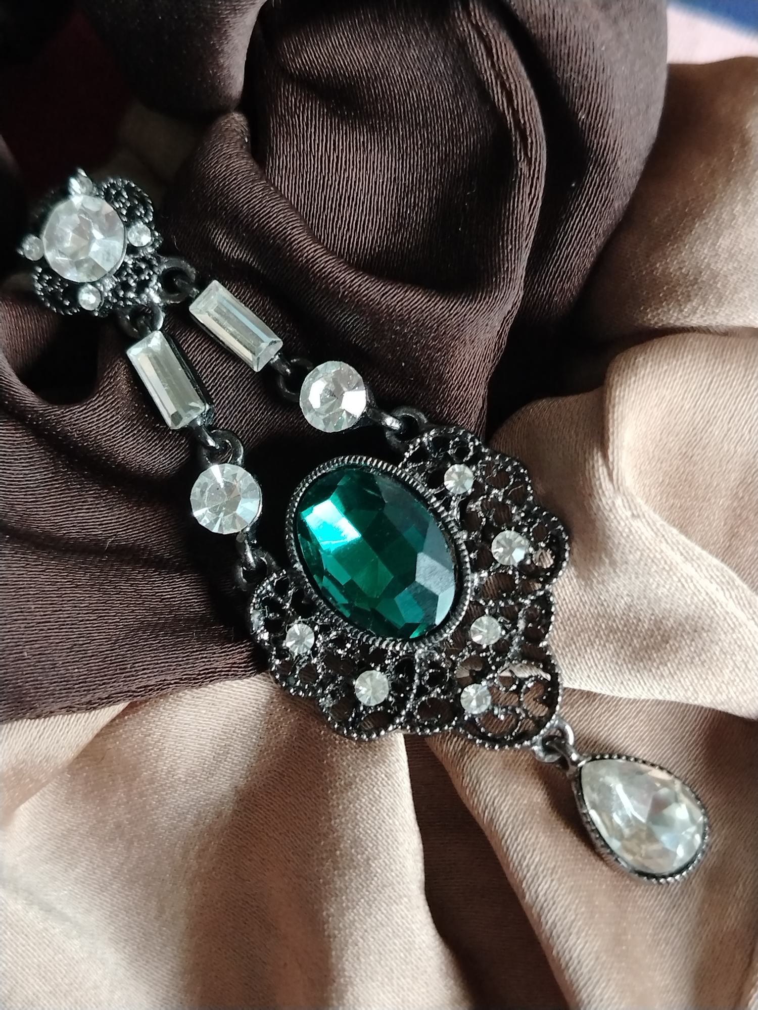 A dark, metallic brooch with a large, faceted emerald-green gemstone is centered on a draped fabric surface. The brooch has an ornate, filigree design around the emerald, studded with small, clear rhinestones. Hanging below the main part of the brooch is a single, clear, teardrop-shaped gemstone. The brooch is attached to a chain that extends upwards and to the left, where it connects to a cluster of clear rhinestones and rectangular clear gemstones, all set in the same dark metal. The fabric beneath the brooch is made up of at least two different materials or colors. On the left and top, a dark brown, satin-like fabric is visible, with visible horizontal stitching or ribbing. To the right and bottom, a lighter, possibly beige or tan, satin-like fabric is draped, with gentle folds and a sheen that catches the light. The lighting appears to be artificial and diffused, as there are no harsh shadows. The focus is entirely on the brooch and its immediate surroundings. No people, text, or specific location cues are visible.