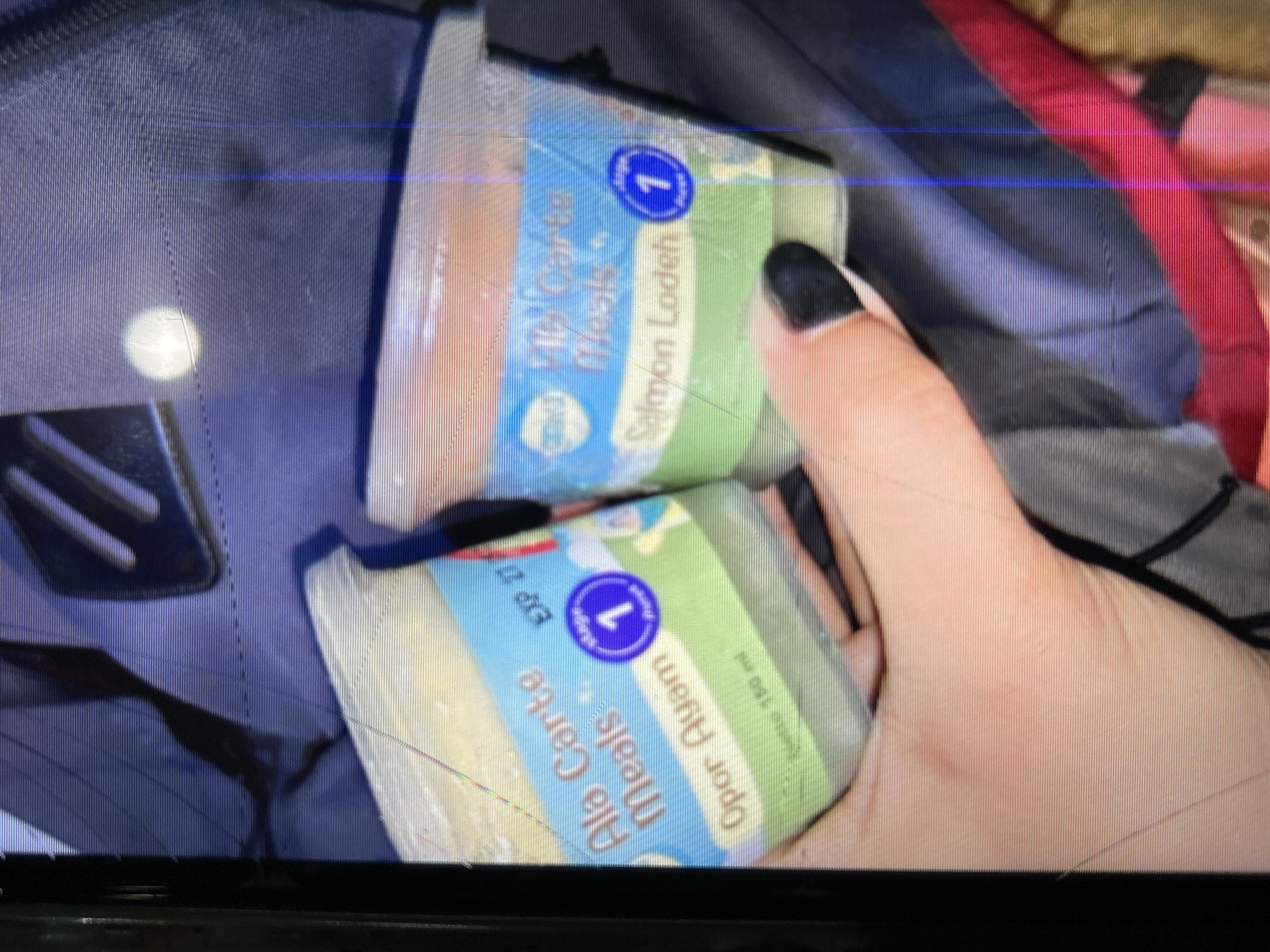 The image shows two small containers of "A La Carte Meals" being held by a person's hand. The left container has text indicating "Opor Ayam" and "EXP 27". The right container shows "Salmon Lodeh". Both containers have a blue circular label with the number "1" inside, suggesting they are for Stage 1. The background appears to be a dark fabric, possibly a backpack. There are visible digital artifacts and lines across the image, indicating a low-quality digital capture. The scene is not identifiable as being in Depok, Indonesia, as there are no visible landmarks or contextual clues to support this location.