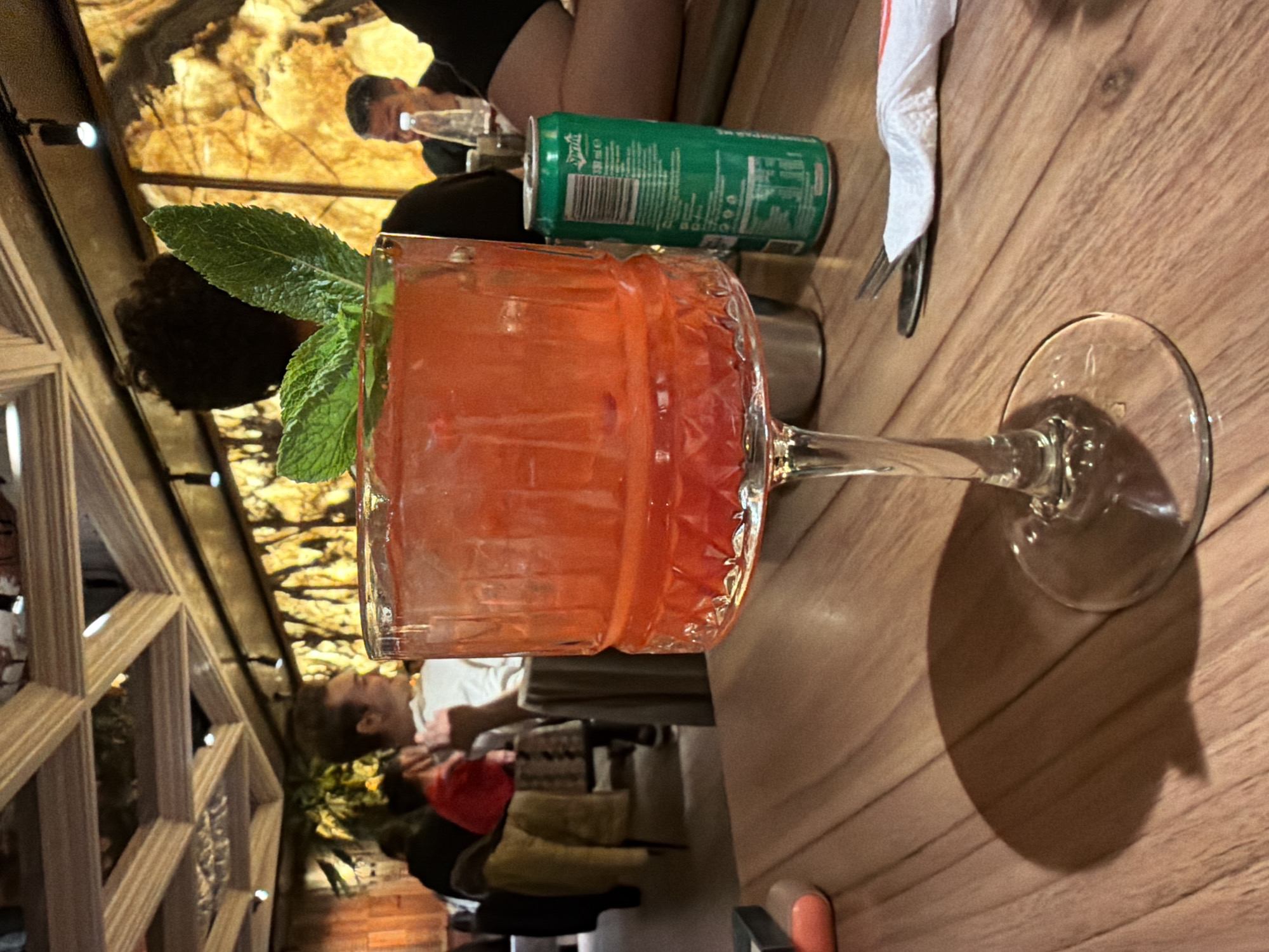 The image shows a cocktail glass filled with a red drink, garnished with mint leaves, on a wooden table in what appears to be a restaurant or bar. Next to the glass is a can of Sprite. The background shows other patrons and some architectural details, including a decorative wooden frame and a wall with a nature scene. Based on the style, the location is likely in Sofia, Bulgaria. There's a fork and folded napkin. People are seated and talking in the background.