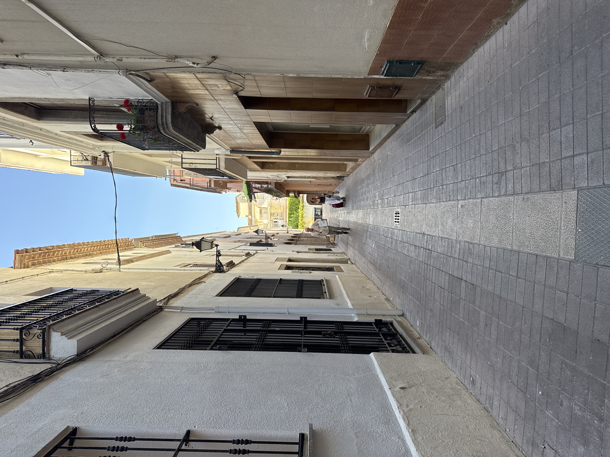 The image captures a narrow, European-style street in Tossa De Mar, Spain. The perspective is low, looking up and down the street, which is paved with gray square tiles. Buildings line both sides of the street, with their facades made of stucco and stone in varying shades of white and beige. Some buildings have balconies with decorative wrought-iron railings, one of which holds a pot of red flowers. Windows are visible, many with black wrought-iron grates.
Two people are visible in the distance, walking away from the camera. One person appears to be wearing a white shirt and dark pants, carrying a red bag. Another person is walking further down the street. A vintage-style street lamp is mounted on the side of one building. The sky is a clear, bright blue, suggesting a sunny day. The shadows cast by the buildings indicate it is likely midday. The overall impression is one of a quiet, picturesque European town.