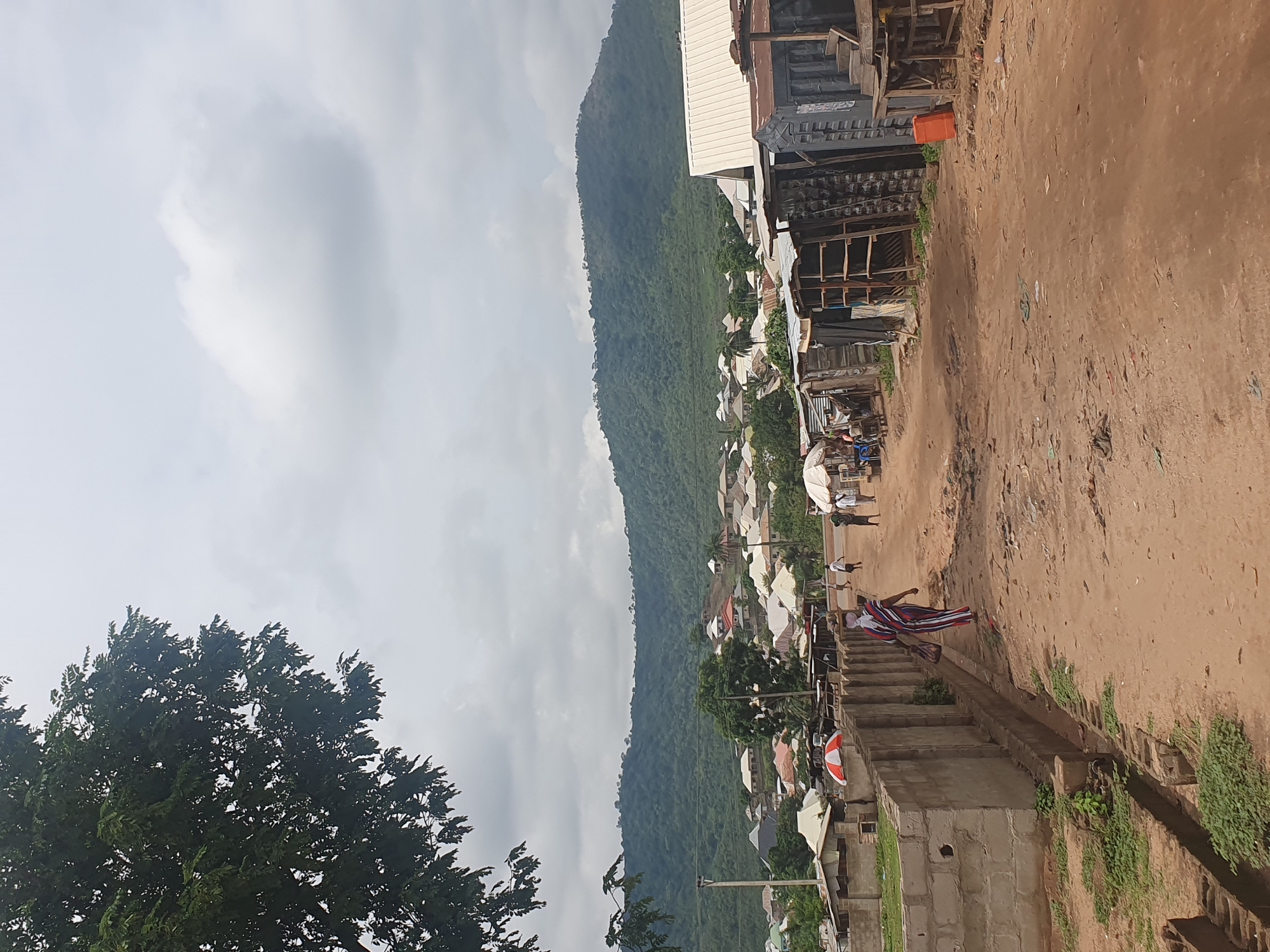 The image shows an elevated view of a street scene in Lokoja, Nigeria. A tree occupies the lower left corner of the frame. The street is unpaved and runs uphill. A mountain ridge is visible in the background, with buildings and houses nestled along its base. Along the street, there are makeshift structures and stalls, suggesting a market or commercial area.
Several people are visible on the street, including one person descending a set of concrete steps on the right side of the image, wearing what appears to be a striped garment. Other individuals are scattered along the street further uphill.
The sky is overcast with clouds. The overall environment appears to be a densely populated area within the city.