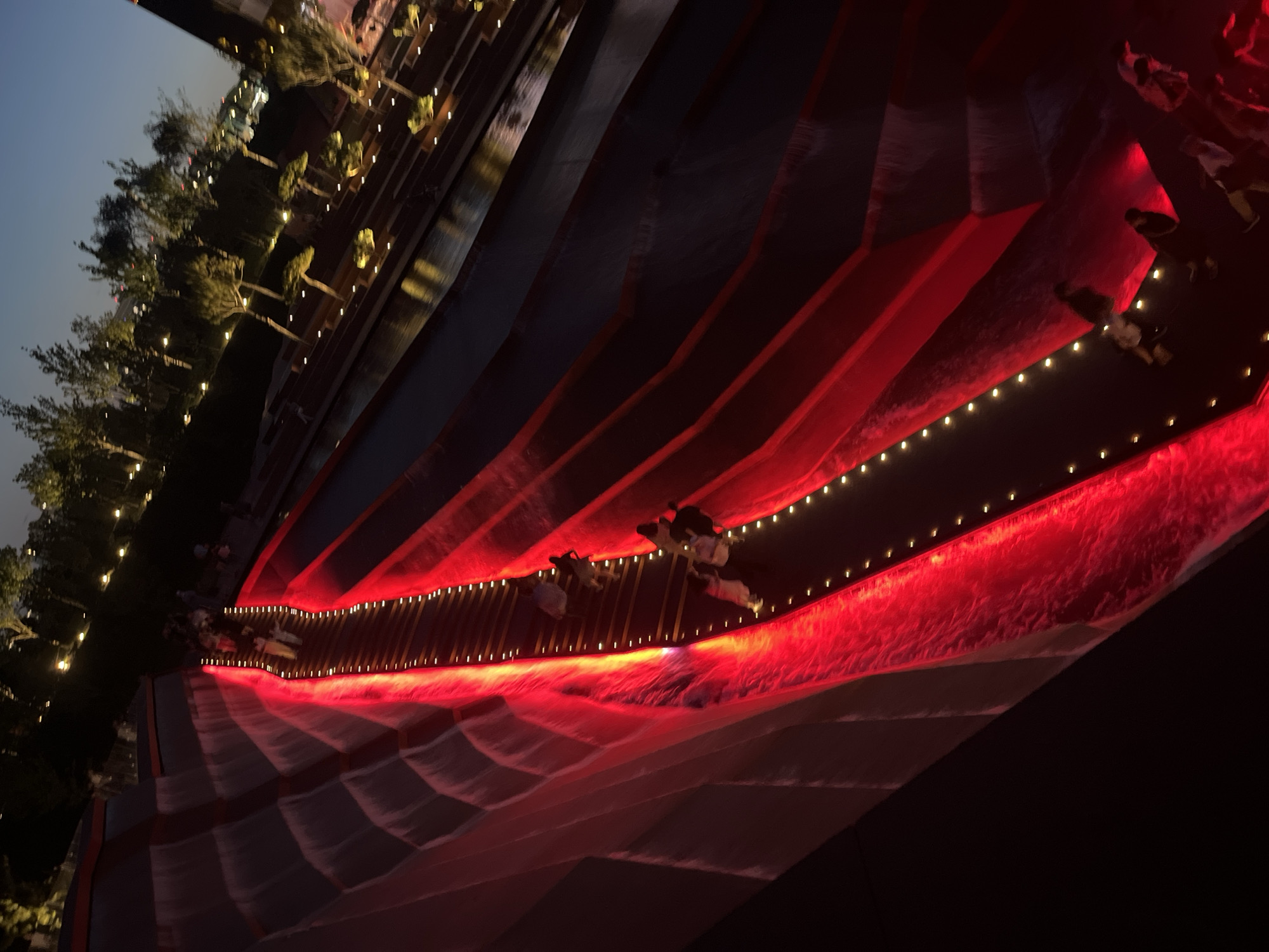 The image shows a nighttime scene in a modern urban park setting. A prominent feature is a large, tiered water feature illuminated with bright red lights. People are walking up a staircase that runs alongside the water feature. The stairs are lined with small, warm-toned lights. Above and to the right of the water feature, there are trees and more lights, suggesting a landscaped area with pathways and potentially buildings in the background. The sky is dark, indicating it is either dusk or nighttime. The overall impression is one of a contemporary, illuminated public space designed for recreation or viewing. This scene is likely located in the city of Krasnodar, Russia, specifically at the "Waterfalls" park.