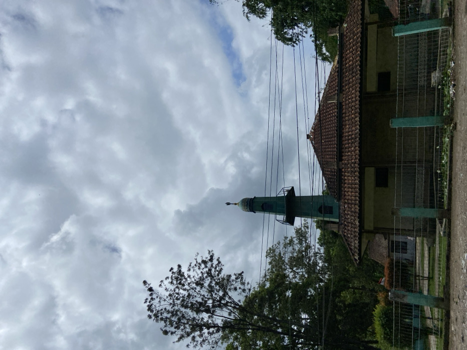 The image shows a mosque in Cipatat, Indonesia, located at coordinates -6.796 latitude and 107.442 longitude. The mosque has a green minaret with a small balcony, a tiled roof, and white walls. The mosque is surrounded by a chain-link fence, and there are trees and power lines in the background. The sky is overcast with white and gray clouds. There is a dirt road in the foreground, and there are some pieces of white trash scattered on the ground.