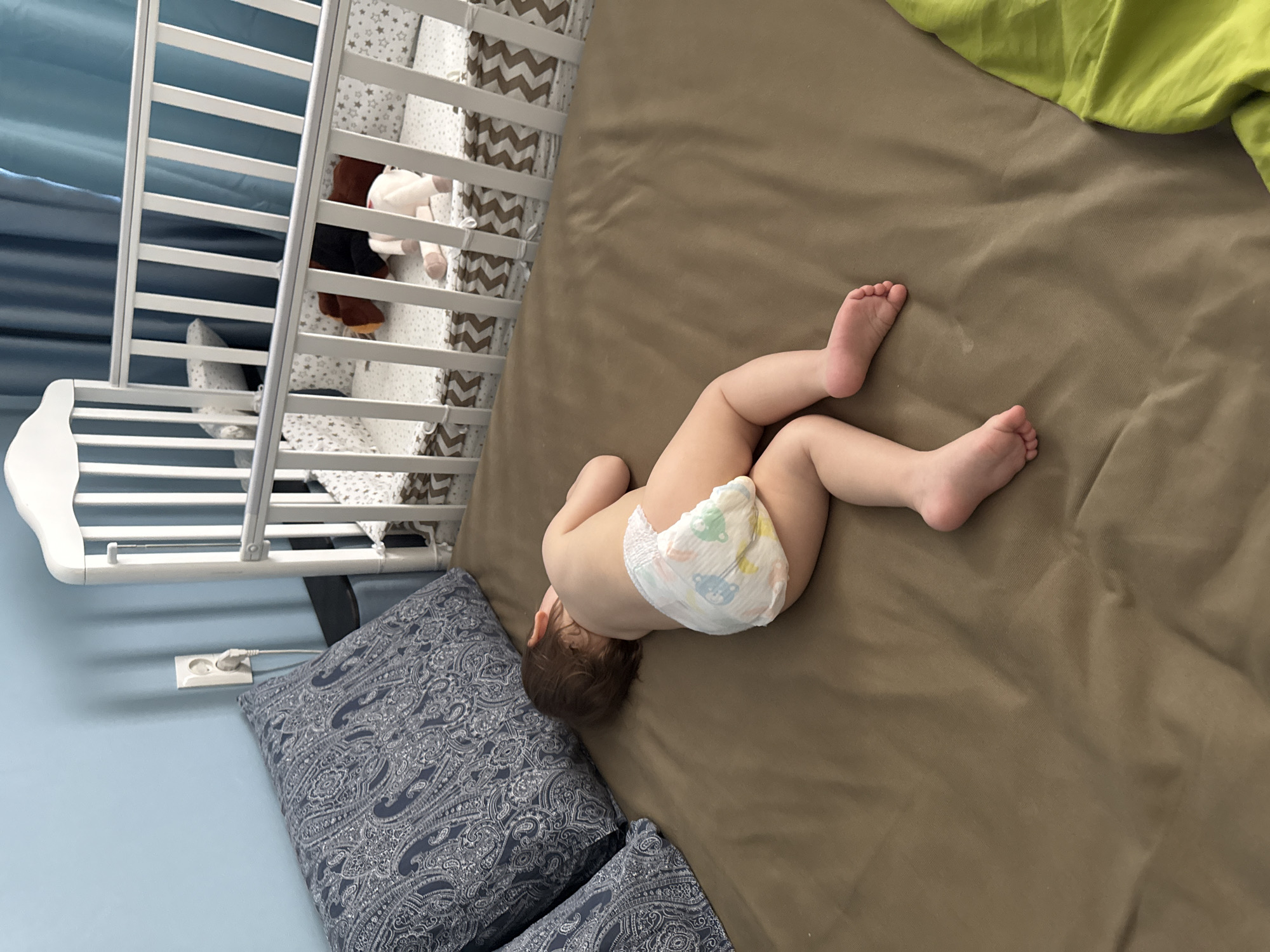 A baby wearing a diaper is lying on a brown sheet next to a crib in a room with light blue walls. There are two pillows with a paisley pattern next to the baby. A section of green fabric is visible in the upper right corner. A white electrical outlet is visible on the wall below the crib. There is no clear indication of the image's location in the city of Moscow, Russia.