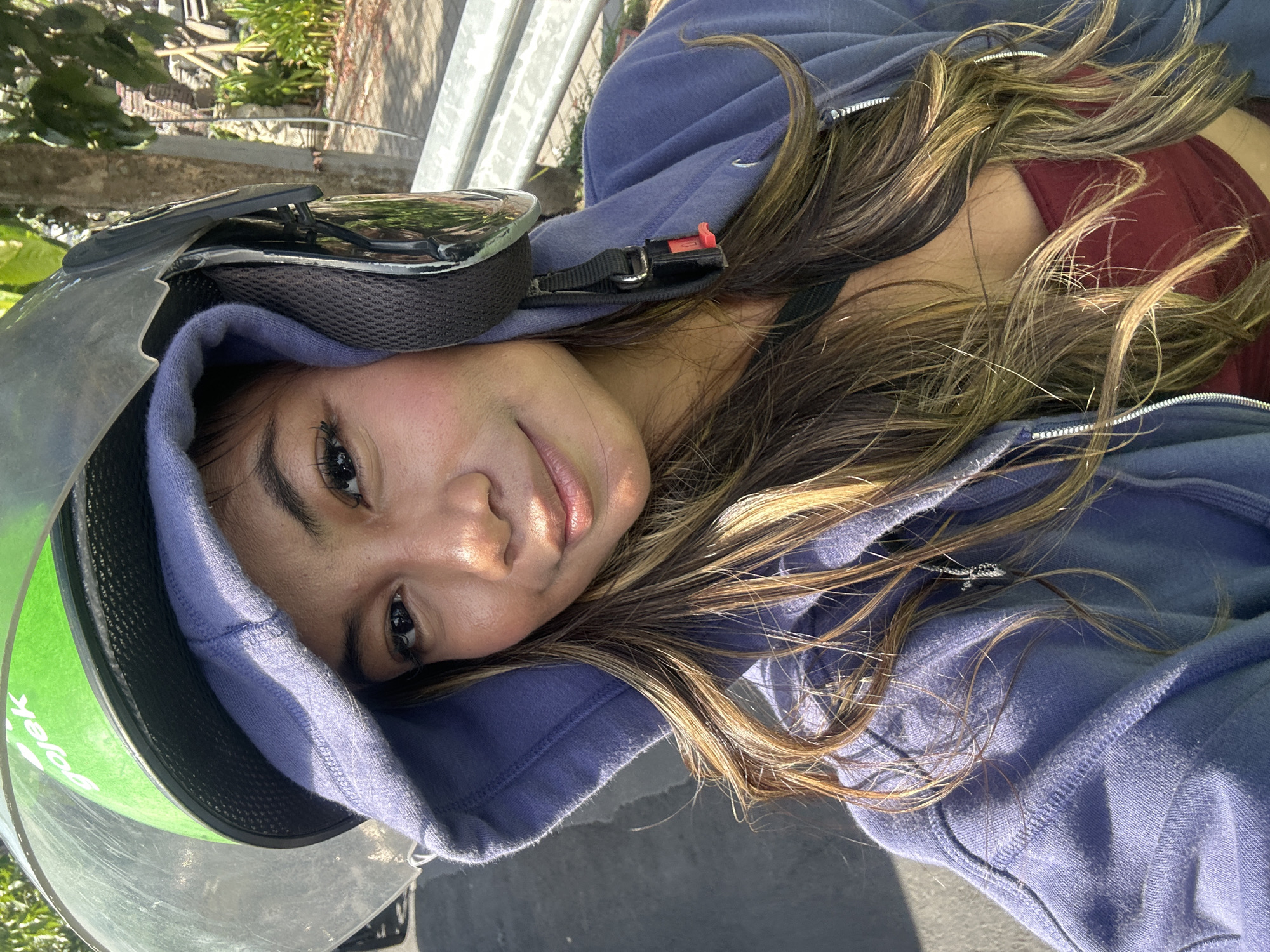 A close-up, outdoor selfie shows a young woman wearing a blue hooded sweatshirt and a green and black motorcycle helmet with a clear visor. The helmet has the word "gojek" in white lettering on the green portion. Her face is centered in the frame, and she is looking at the camera with a slight smile. Her long, brown, highlighted hair is visible around her face and shoulders. She is wearing a dark red camisole underneath her sweatshirt. The background suggests an urban environment with a paved road, some greenery, and a light-colored railing. The lighting indicates it is daytime, and the shadows suggest sunlight. The scene is likely taking place on a street in Denpasar, Indonesia, given the presence of "Gojek," a prominent ride-hailing and delivery service in Indonesia.