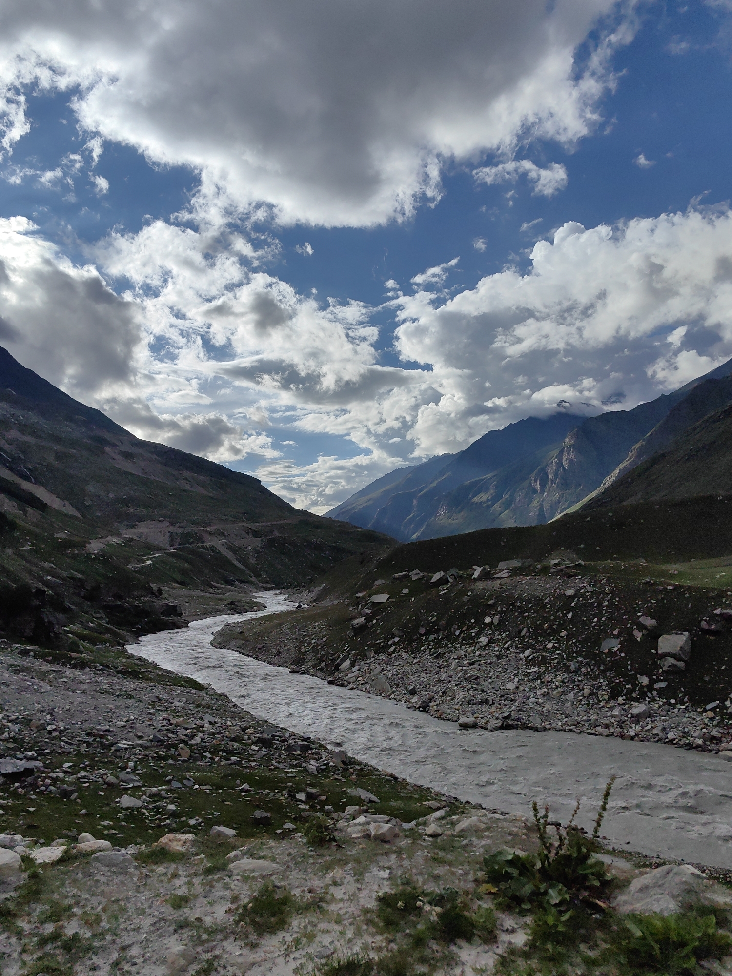 A wide, muddy river flows through a valley surrounded by rugged, green mountains under a dramatic sky filled with dark, ominous clouds and patches of bright blue. The river curves from the middle distance towards the foreground, its surface disturbed by ripples and small waves. The valley walls are steep and covered in sparse vegetation and rocky scree. A winding dirt road is visible on the left side of the valley, carved into the mountainside. The foreground features rough, rocky terrain with some tufts of green grass. The scene appears to be a natural landscape in a remote, mountainous region, possibly in the Himalayas, given the reference to Burwa, India, which is a village located in the Ladakh region. There are no people or man-made structures other than the road.