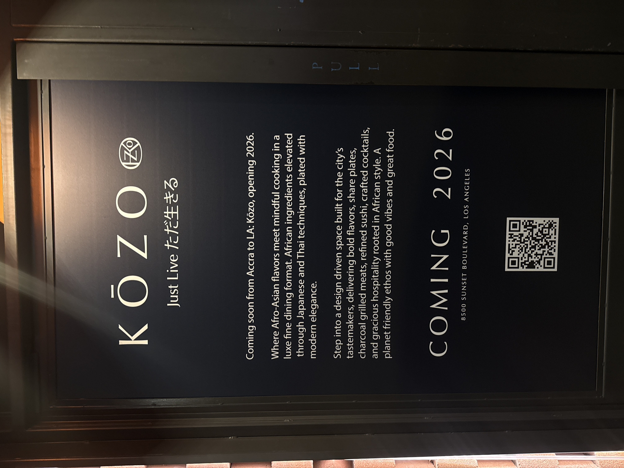 The image displays a vertically oriented, dark-colored sign or poster, likely mounted within a frame or part of a building facade, announcing the upcoming opening of a restaurant. The sign features white text against a dark background, with the prominent restaurant name "KŌZO" and the tagline "Just Live ただ生きる" (Just Live).
The sign details that Kōzo is "Coming soon from Accra to LA," with an opening projected for 2026. It describes the establishment as offering "Afro-Asian flavors" through "mindful cooking in a luxe fine dining format," blending African ingredients with Japanese and Thai techniques. The space is touted as "design driven" for "the city's tastemakers," promising "bold flavors, share plates, charcoal grilled meats, refined sushi, crafted cocktails, and gracious hospitality rooted in African style," all adhering to a "planet friendly ethos with good vibes and great food."
At the bottom right, a QR code is visible, next to the full address: "8500 SUNSET BOULEVARD, LOS ANGELES." Faintly visible at the top, on what appears to be a door or window frame, is the word "PULL." The image is taken at an angle, suggesting an upward view or a tilted perspective. The setting appears to be a building exterior or an indoor display within the West Hollywood, United States of America area, as indicated by the Sunset Boulevard address. There are no people or specific activities depicted, only the informational sign.