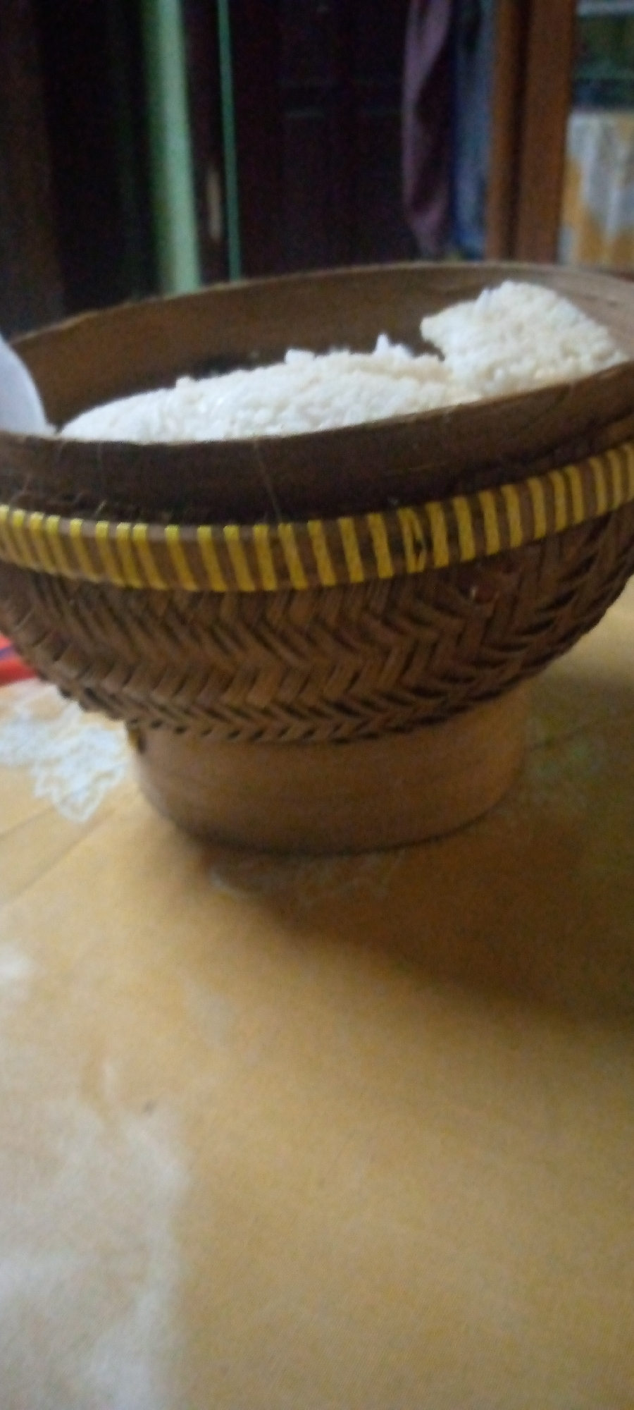 A woven basket filled with white rice sits on a yellow surface. The basket has a woven pattern and a band of yellow and brown stripes around the rim. The background is dark and out of focus, with hints of vertical lines and fabric. The overall scene suggests a domestic setting in Sumberwono, Indonesia.