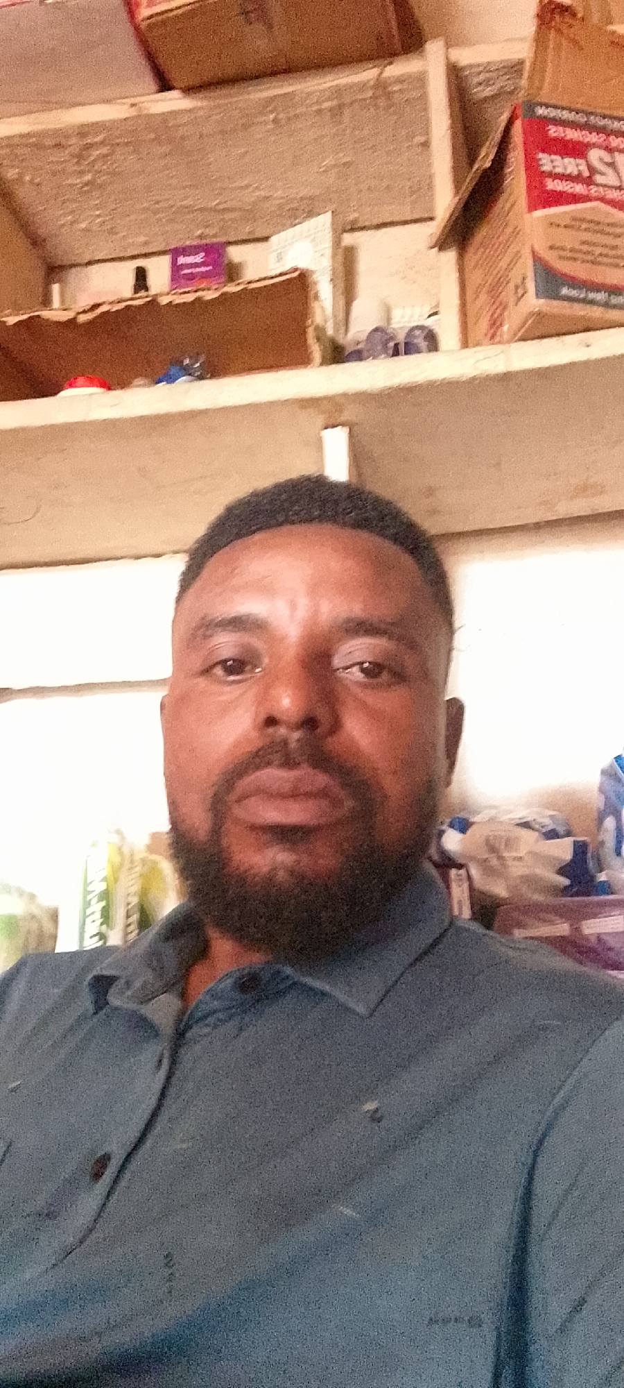 A medium shot portrait photograph shows a Black man with dark skin, a short haircut, a beard, and a mustache. He is wearing a dark blue collared shirt. He is looking directly at the camera. The background is out of focus, but appears to be a room with shelves filled with various items, including boxes and what appear to be products. The lighting is dim and appears to be coming from the front. The photograph was taken in Durumi, Nigeria.