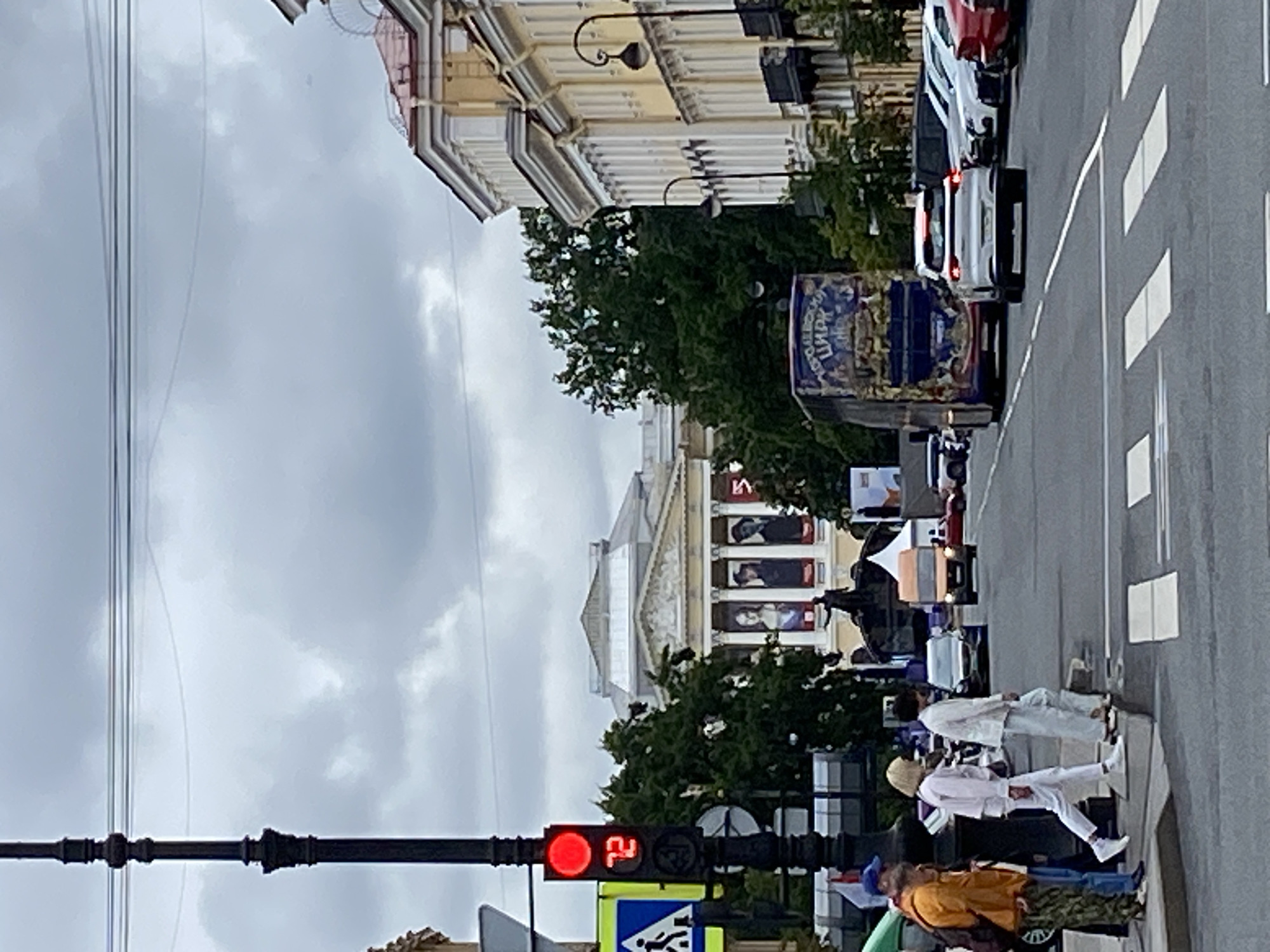 This image captures a street scene in Saint Petersburg, Russia, likely near a cultural institution given the architectural style of the buildings and the presence of large banners on the facade of the prominent building in the background, which resembles the Alexandrinsky Theatre.
In the foreground, a traffic light displays a red signal and the number "72", indicating the pedestrian crossing signal is active. Several individuals are crossing the street, with some waiting at the curb. The group includes a person in an orange jacket, another in a white shirt and pants, and a third in a white suit. They are on a paved pedestrian crossing.
The street itself is asphalt with white lane markings and a crosswalk. Cars are present, including a silver SUV in the middle lane, a large double-decker bus with colorful advertisements on the side (one banner reads "КОРОЛЕВСКИЙ ЦИРК", meaning "Royal Circus"), and several parked vehicles, including white sedans and a red car, on the right side of the road.
The environment is urban, with tall, classical-style buildings lining the street. Trees with green foliage are visible behind the crosswalk, and overhead power lines are strung across the sky. The sky is overcast, suggesting cloudy weather. A lamppost with an ornate design stands on the left side of the image. A pedestrian crossing sign with a silhouette of a person is also visible near the traffic light.