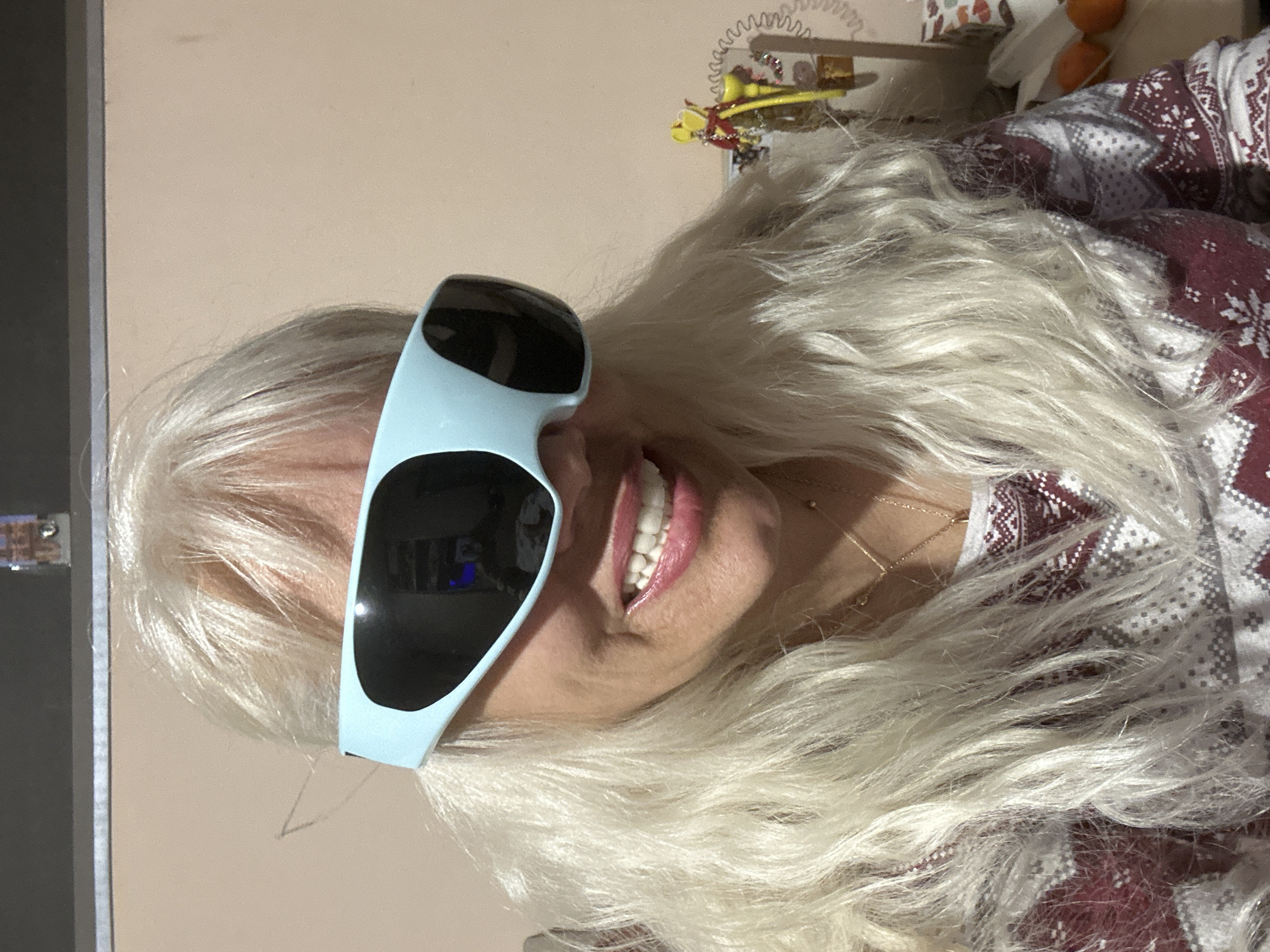 A woman with white-blonde, shoulder-length hair is smiling and wearing oversized, light-blue sunglasses with dark lenses. She is wearing a maroon and white patterned shirt or sweater. A thin gold necklace is visible around her neck. Behind her, on a light peach-colored wall, there is a telephone receiver, various decorative objects, and some oranges. A dark gray wall or door frame is visible in the left side of the image. The location cannot be determined as being in Sofia, Bulgaria, from the information available.