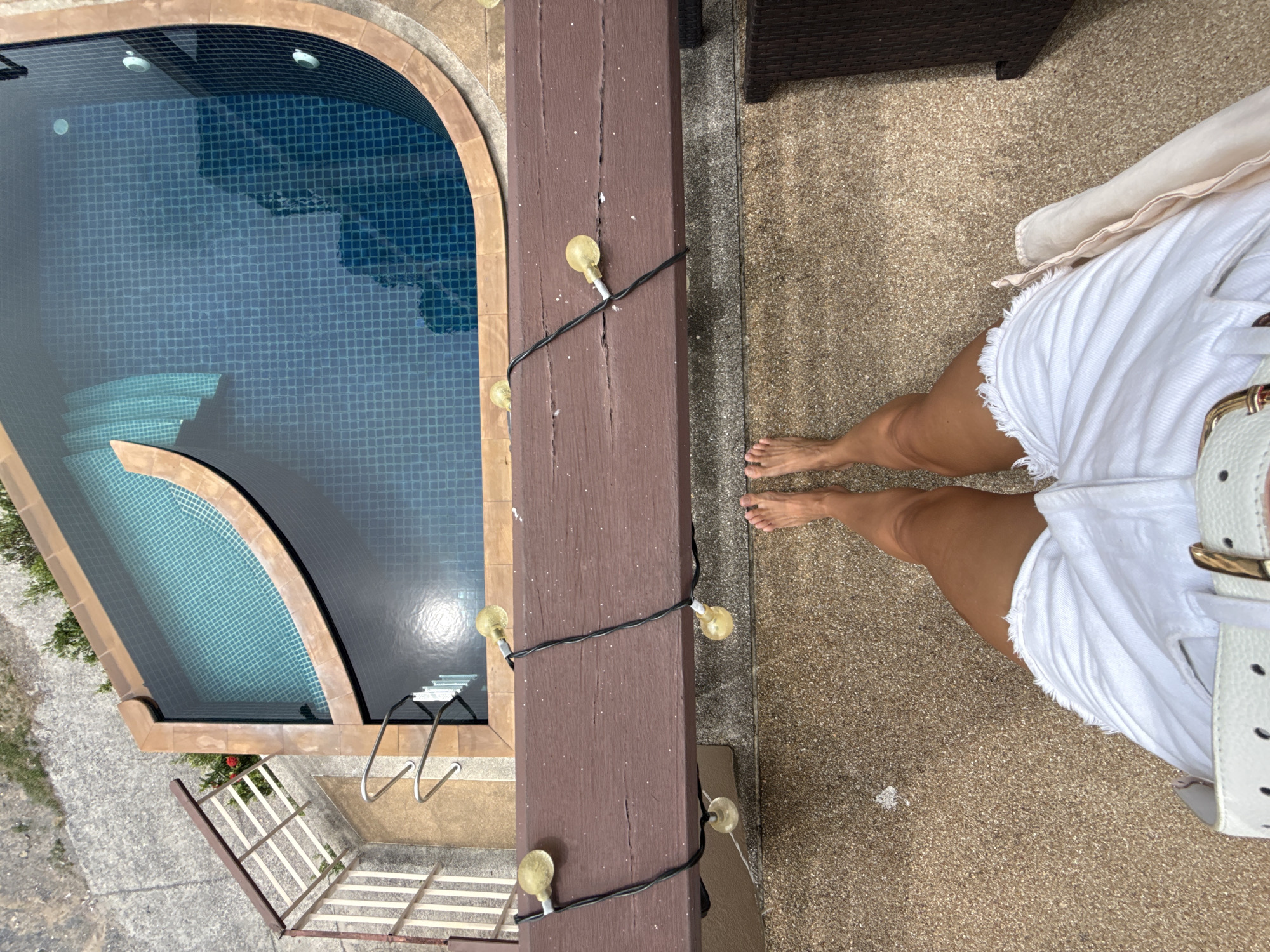 An overhead, eye-level shot features a woman's bare feet and white denim shorts on a gravel-like patio. Adjacent to the patio is a wooden railing adorned with a string of yellow globe lights. Beyond the railing lies a blue tiled swimming pool with a curved edge, reflecting sunlight on its surface. The pool area is bordered by tan tiles and a concrete pathway, with a glimpse of green foliage and a railing on the far left. The scene appears to be set in a residential outdoor area, possibly in Baan Chaweng, Thailand.