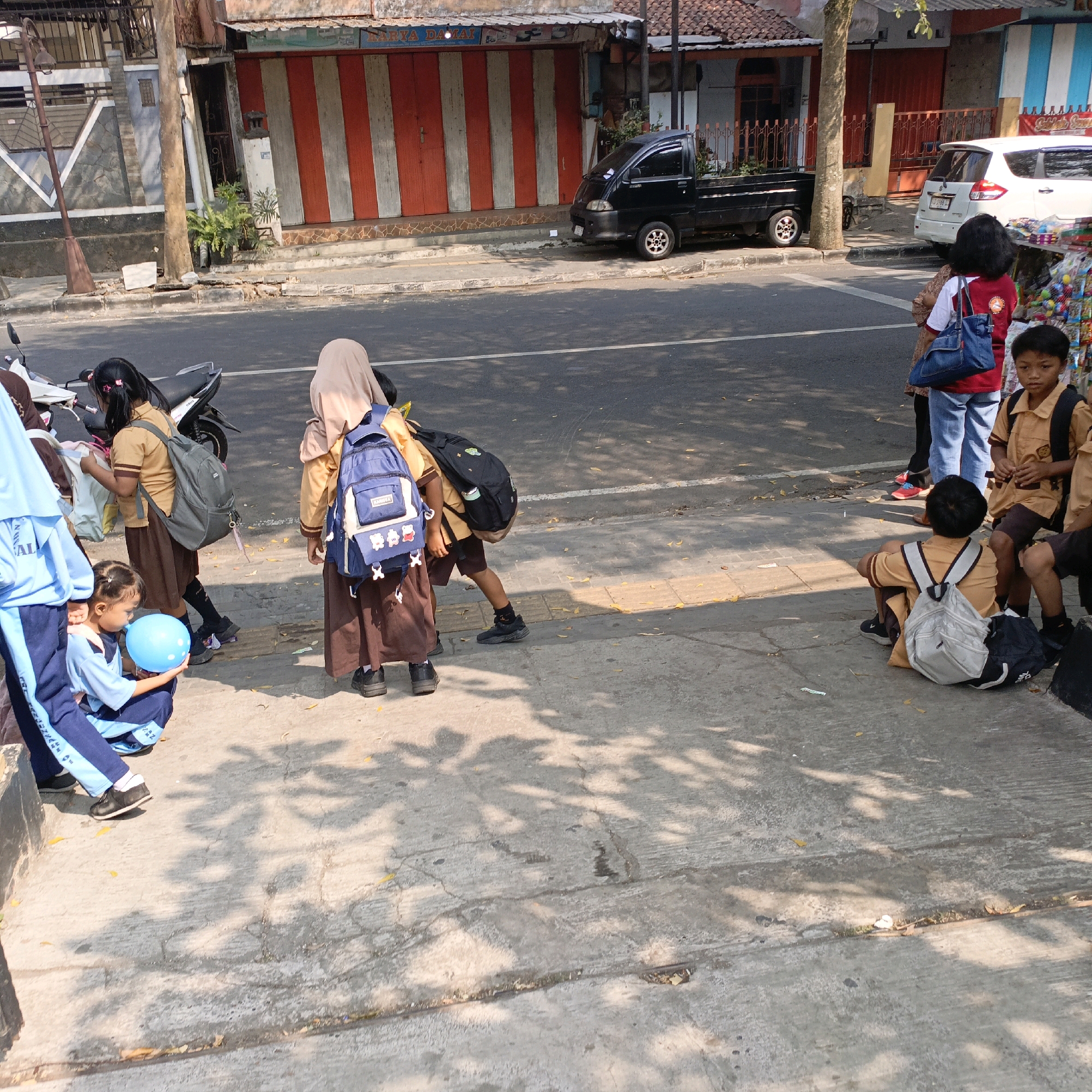 The image captures a sunny daytime scene on a street in Salatiga, Indonesia, likely in front of shops. Several school children, dressed in uniforms, are visible. One child in a light blue uniform is crouching and holding a blue balloon. Another child, in a tan uniform and hijab, stands with a blue backpack. Other children in similar uniforms are also present, some with backpacks, suggesting they are on their way to or from school. The background shows a row of shops with red and white striped roll-up doors and other architectural details typical of Indonesian streetscapes. A black pickup truck is parked near the shops, and a white car is further down the street. The sidewalk is paved and partially covered in shadows cast by overhead trees. A motorcycle is parked on the left side of the image. The overall impression is one of a typical street scene in a bustling Indonesian city during daylight hours.
