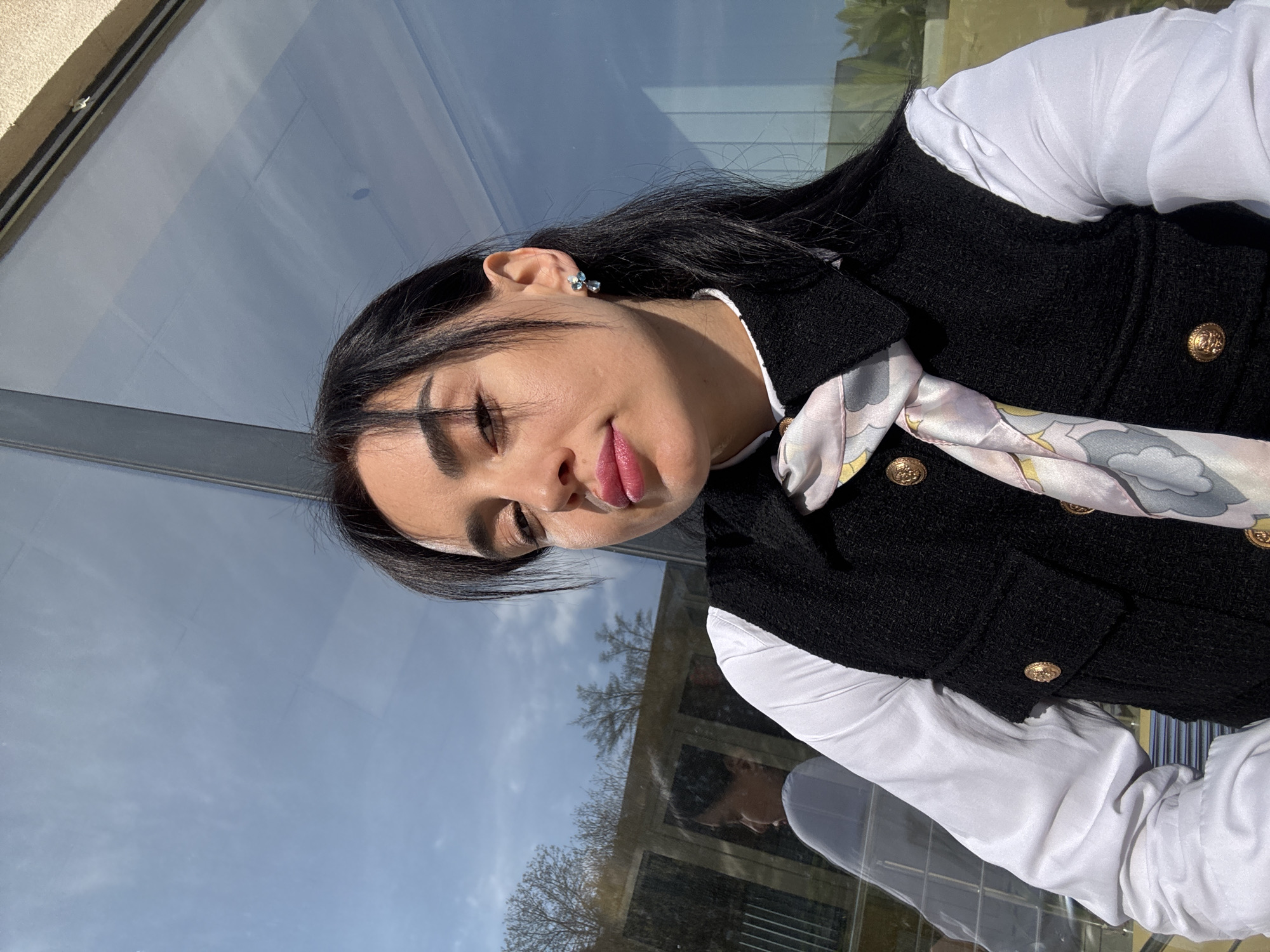 A portrait photograph captures a young woman positioned indoors, appearing to be next to a large glass window or wall. The image is rotated, showing her head tilted, with a soft, engaging expression, dark eye makeup, and pink lipstick. She has long, dark hair styled down, and a small, light blue earring adorns her left ear.
She is dressed in a crisp white long-sleeved shirt, over which she wears a textured black sleeveless vest or jacket. The vest features several ornate, gold-toned buttons on the front and pocket flaps. A light-colored silk scarf with an abstract pattern in shades of pink, blue, yellow, and white is visible, tied around her neck and tucked into the vest.
The background is dominated by reflections in the glass surface. A bright, clear blue sky with a few faint clouds is prominently reflected, indicating a sunny daytime setting. Bare tree branches are also visible in the reflections, suggesting a season without full foliage. Architectural elements, possibly from surrounding buildings or the structure she is in, are subtly reflected, along with a blurred reflection of another individual in a white shirt, likely inside. The overall ambiance suggests a bright day in an urban or modern architectural setting in Tbilisi, Georgia. There is no visible text in the image.
