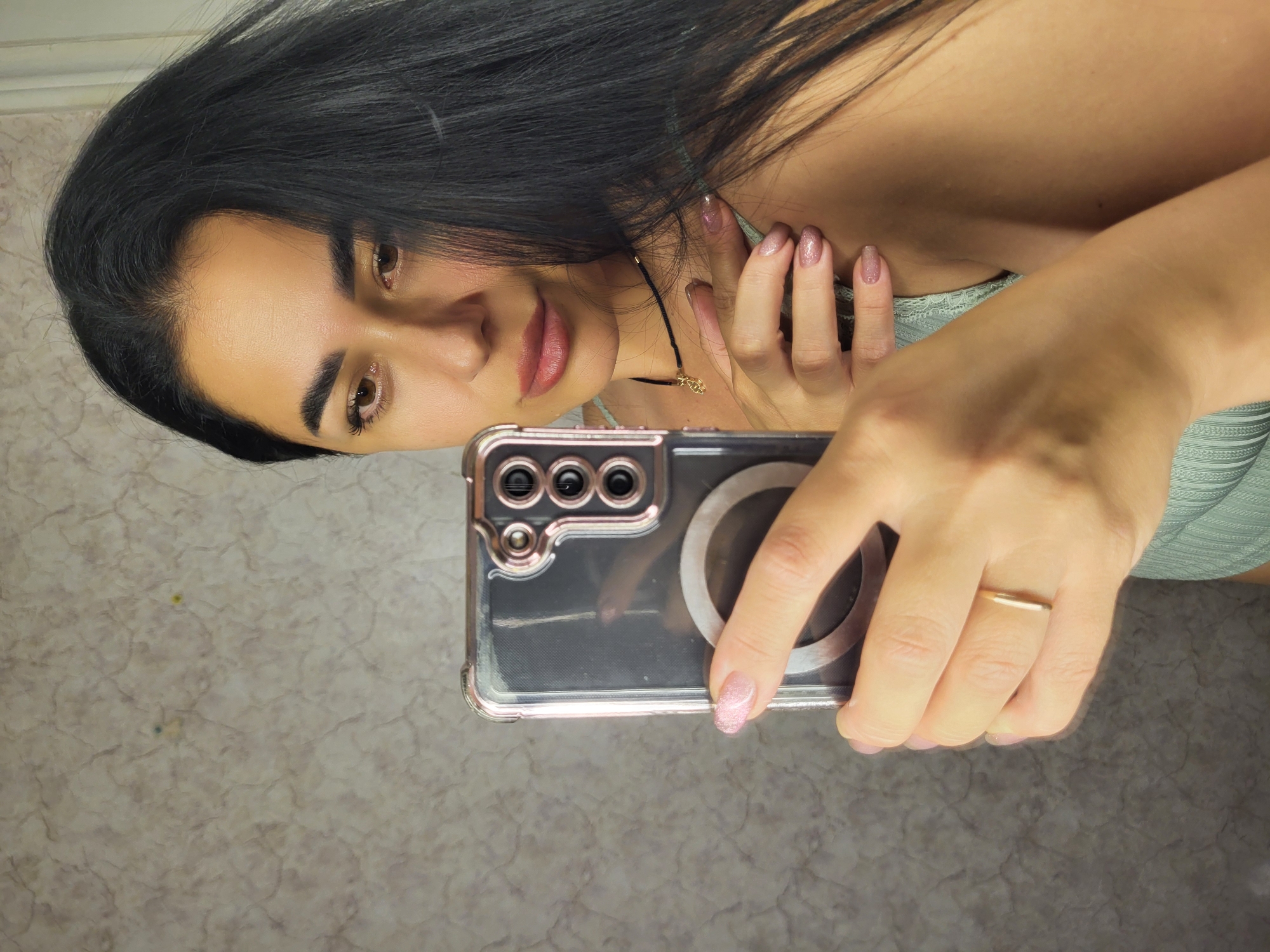 The image shows a young woman taking a selfie with her phone. She is holding the phone in her right hand, and her left hand is resting on her shoulder. She has long, dark hair and is wearing a light green top. The background is a textured wall. The image appears to have been taken indoors, possibly in a home or hotel room. There are no indications of the specific location within Horishni Plavni, Ukraine.
