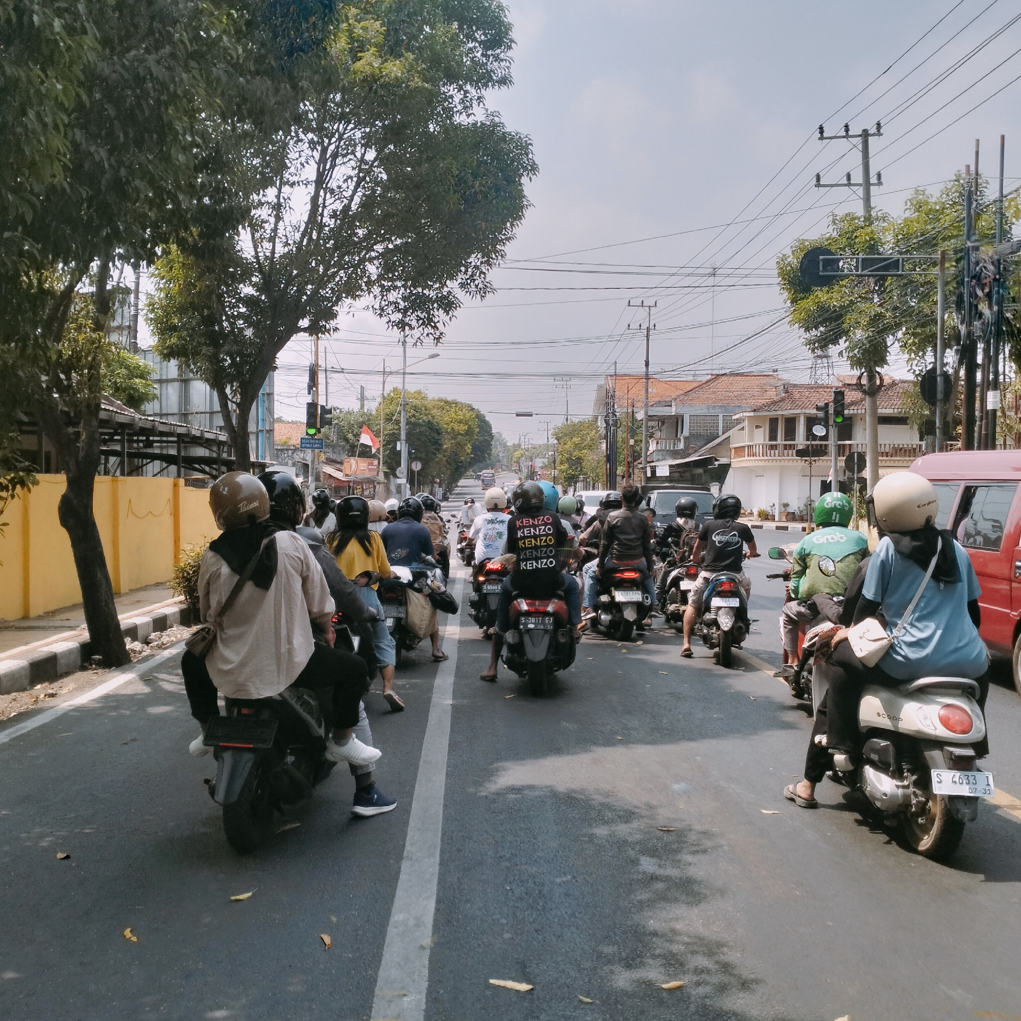 A traffic scene unfolds in Semanding, Indonesia, as a group of motorcyclists navigates a road lined with trees and buildings. The sky is overcast, casting a diffused light over the scene. Several motorcyclists, wearing helmets, are visible in the foreground and mid-ground. One rider in the foreground wears a light-colored shirt and black pants, with a black bag slung over their shoulder. To their right, another rider wears a blue shirt and black pants, with a light-colored bag on their shoulder. Further down the road, more motorcyclists are visible, some with passengers. The road is asphalted and marked with a white line. On the left side of the road, a yellow wall with a sidewalk runs alongside. Trees with green foliage are abundant on both sides of the road. In the background, buildings with tiled roofs and utility poles with overhead wires are present, indicating an urban or semi-urban setting. A traffic light is visible, showing a green signal. The overall impression is one of moderate traffic flow on a typical day in Semanding.