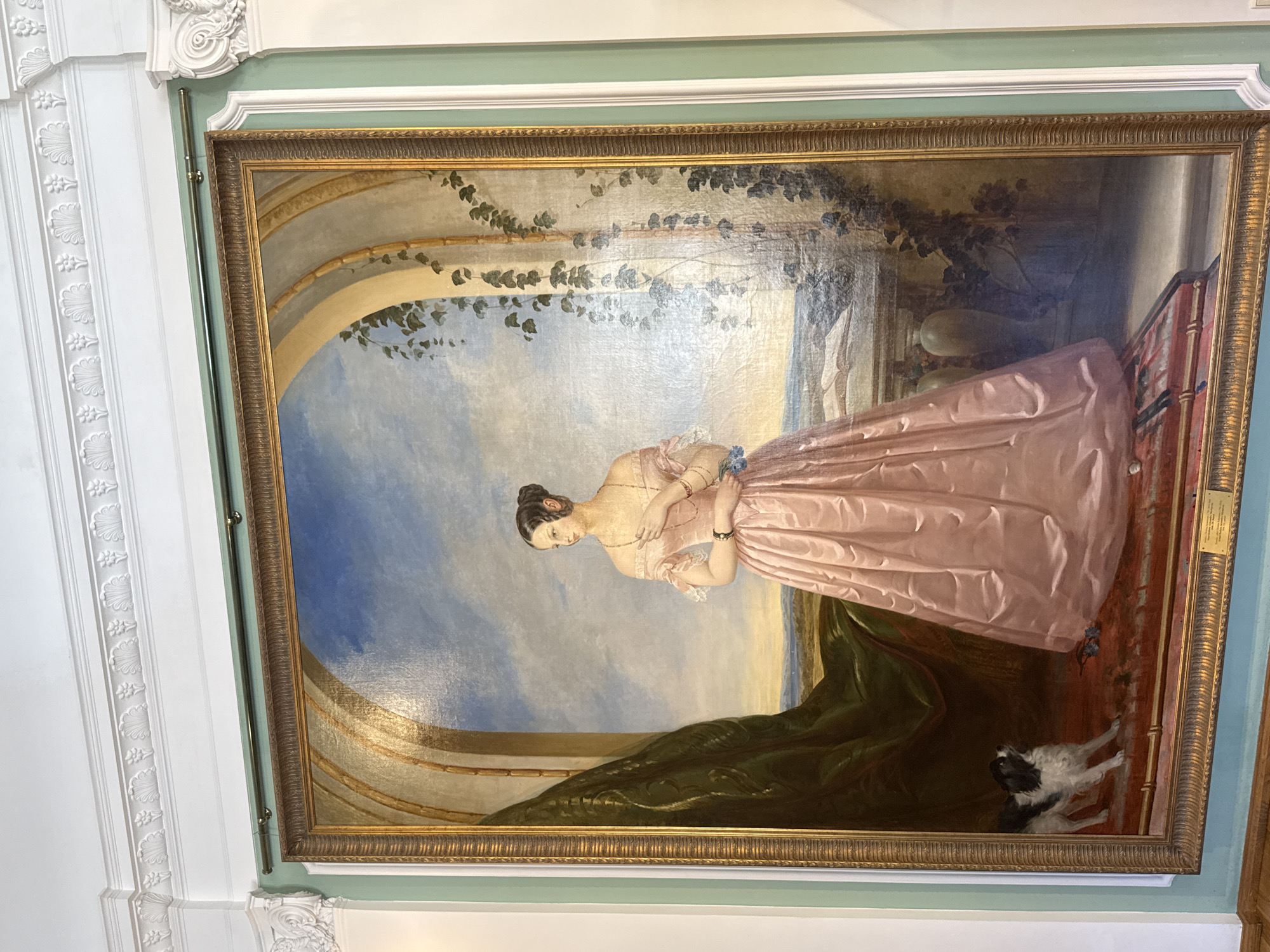 This is a full-length, formal oil painting of a woman in a pink, off-the-shoulder gown. She is standing indoors, likely in a grand room, with an arched architectural element behind her. The background features a landscape with a body of water, mountains, and a cloudy sky.
The woman has dark hair pulled up in a bun and is wearing jewelry, including a necklace and bracelets. She is holding a small bouquet of blue flowers. Her left hand rests on her chest, and her right hand is extended slightly.
In the lower left corner of the painting, a small black and white dog is depicted looking up at the woman. The dog is on a red rug, as is the woman.
The painting is framed by an ornate, gold-colored frame and is hung on a pale green wall. Above the painting, there is white architectural molding with decorative details. The scene is lit from above and to the left, casting subtle shadows. There is a small plaque at the bottom of the painting that is too small to read. The overall impression is one of elegance and refinement.