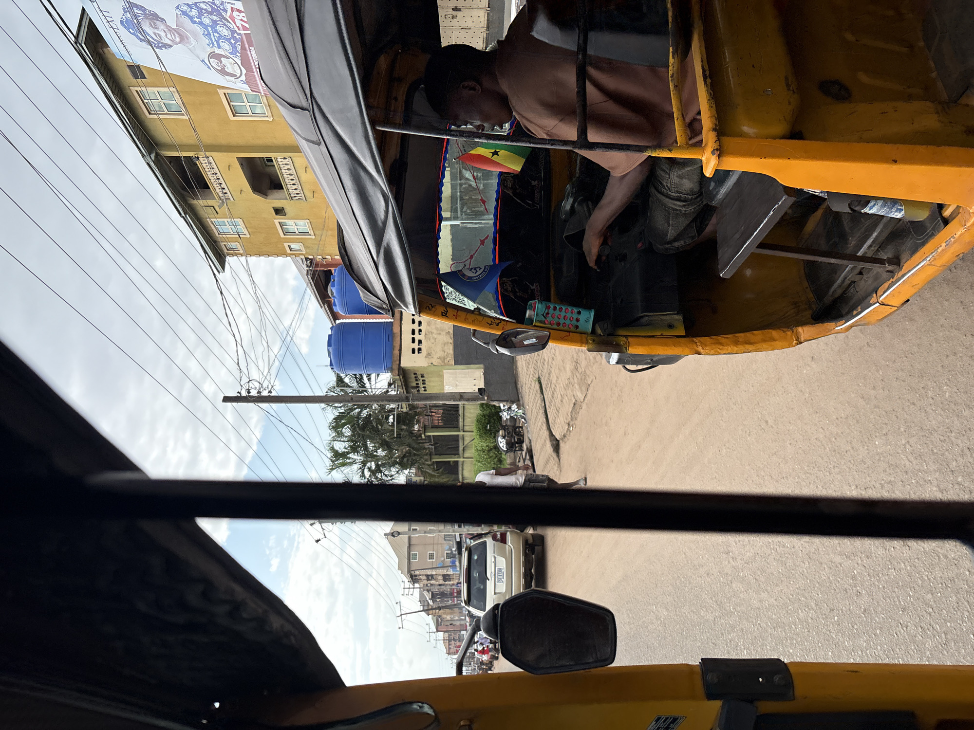 The image, taken from inside a yellow auto-rickshaw, shows a street scene in Nkpor, Nigeria. The driver, a dark-skinned male wearing a brown shirt and dark shorts, is seated at the controls of the auto-rickshaw, looking to his right. A Ghanaian flag and another flag with blue and red accents are visible behind him, attached to the vehicle. The auto-rickshaw is yellow with a black canopy.
Outside the auto-rickshaw, a sandy road stretches ahead, with a silver SUV in front of it. On the right side of the road, a yellow building with multiple windows and balconies stands tall. A large poster featuring a woman's portrait is affixed to the building. Two large blue water tanks are positioned near the building. The sky above is partly cloudy, with visible power lines crisscrossing the frame. Palm trees are sparsely scattered along the street. The overall impression is of a typical urban street in a tropical climate, with vehicles, buildings, and utility infrastructure present.
