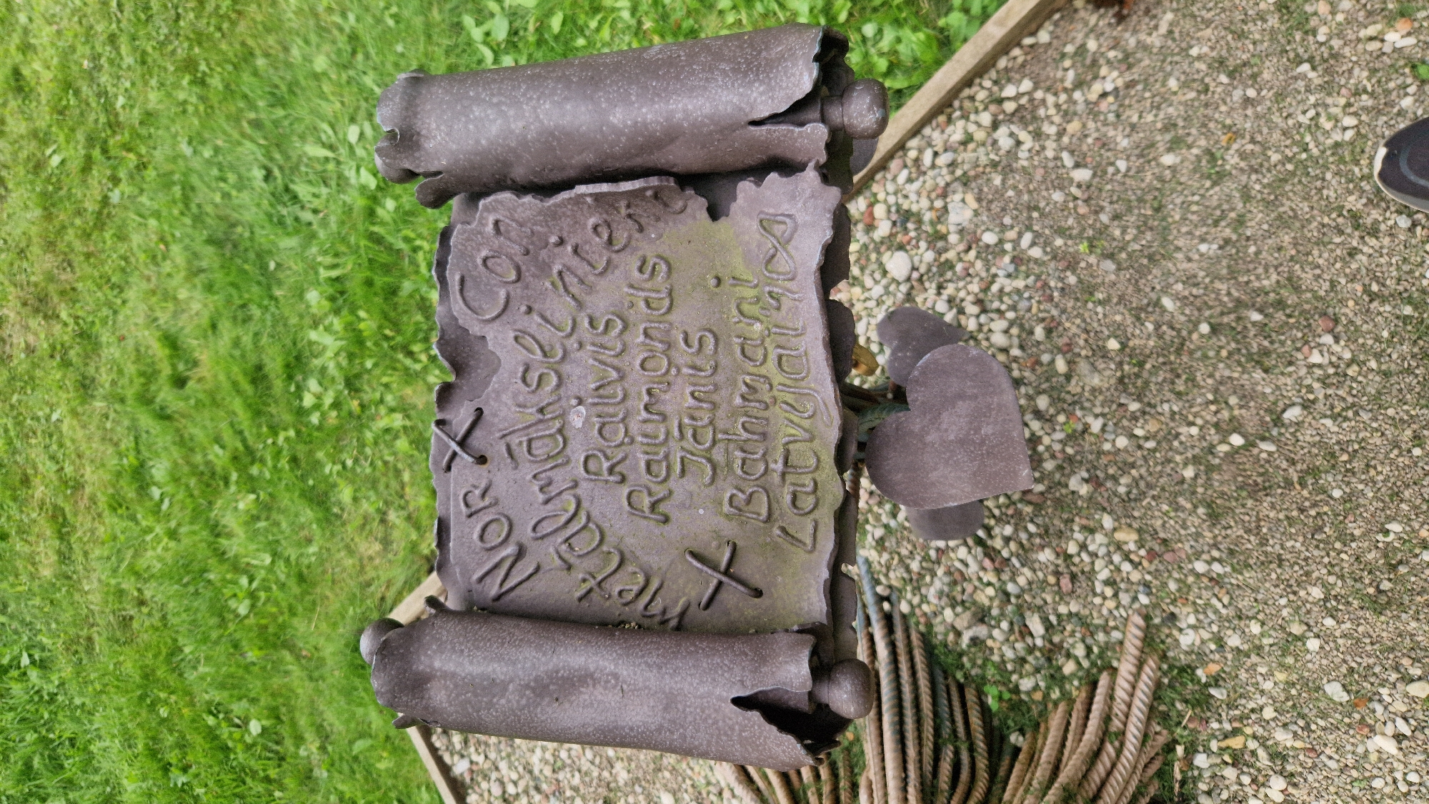 The image shows a metal scroll with names and text inscribed on it. The scroll is made of rusted metal and appears to be part of an outdoor display, possibly a sculpture or memorial. The text on the scroll is in Latvian and reads "NOR metālmāksles", "Raivis", "Raimonds", "Janis", "Bahmani", and "Latvijal". Below the scroll, there are several metal hearts. The background consists of grass and gravel. The scene is likely located in Kazdanga, Latvia, given the reference to "Latvijal" which can be interpreted as "Latvia".