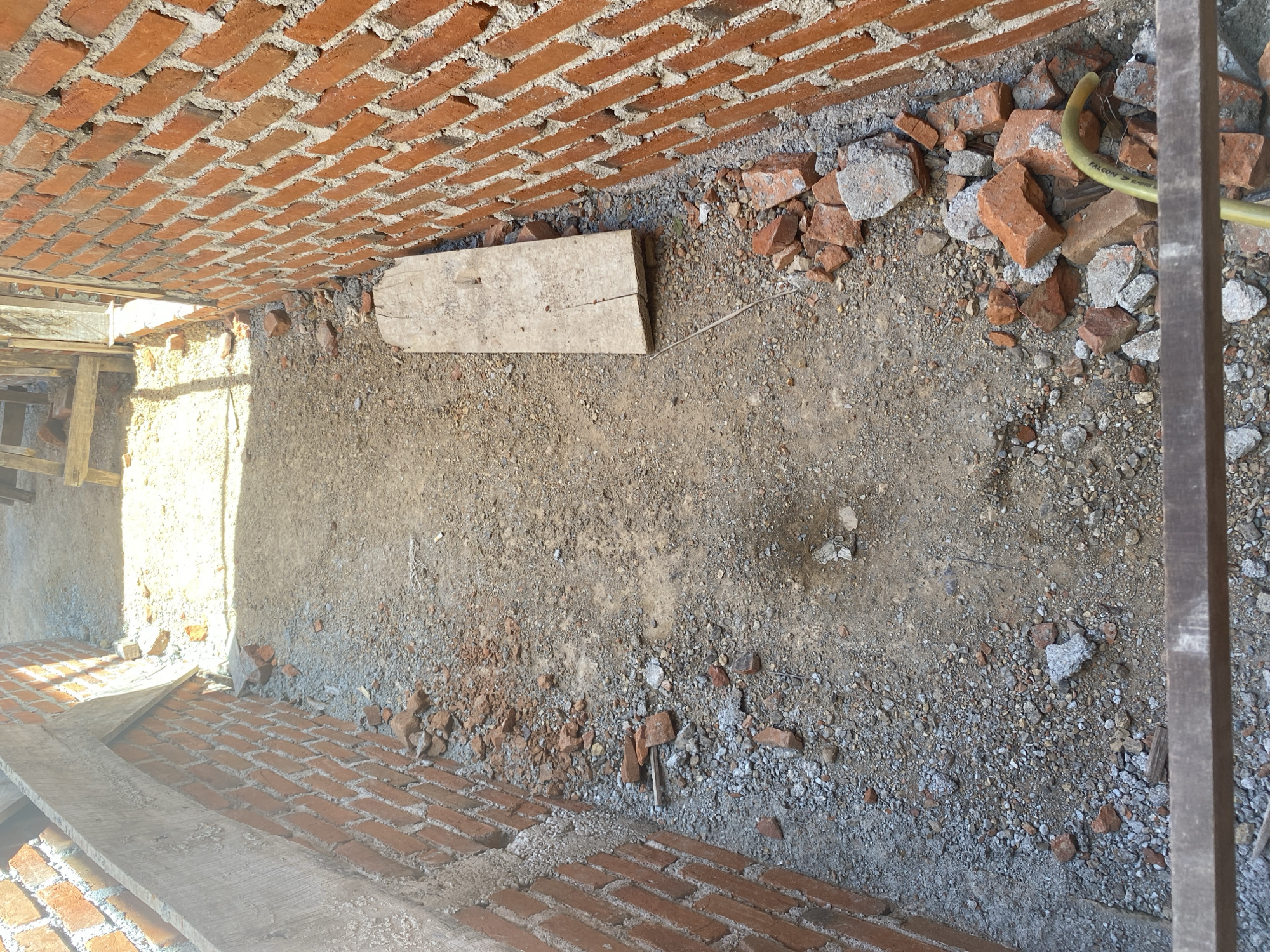The image shows a narrow passage between two brick walls, possibly under construction or renovation, in Banda Aceh, Indonesia. The ground is covered with dirt, gravel, and scattered bricks, indicating a work in progress. Sunlight casts shadows, suggesting it is daytime. There are no people or animals visible in the frame.