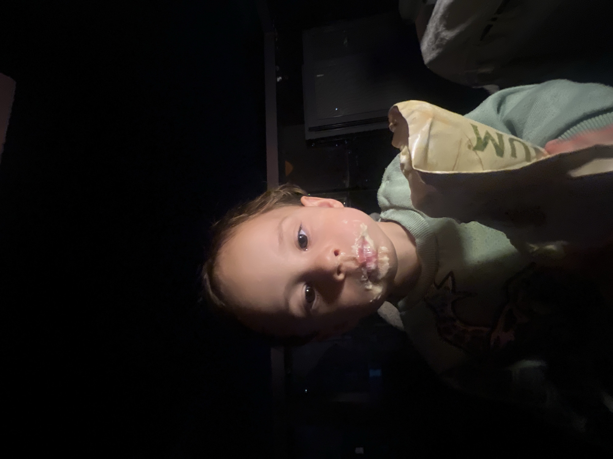 A young child, presumably a Caucasian girl, is holding an item wrapped in paper with the word "WUM" printed on it. The child's face is covered in what appears to be food residue. The environment is dark, with a glimpse of what appears to be an electronic device mounted on the wall. The location is specified as Kiryat Bialik, Israel. The child's clothing is a light green sweater with a graphic image on it. The action is eating or having eaten from the held item.