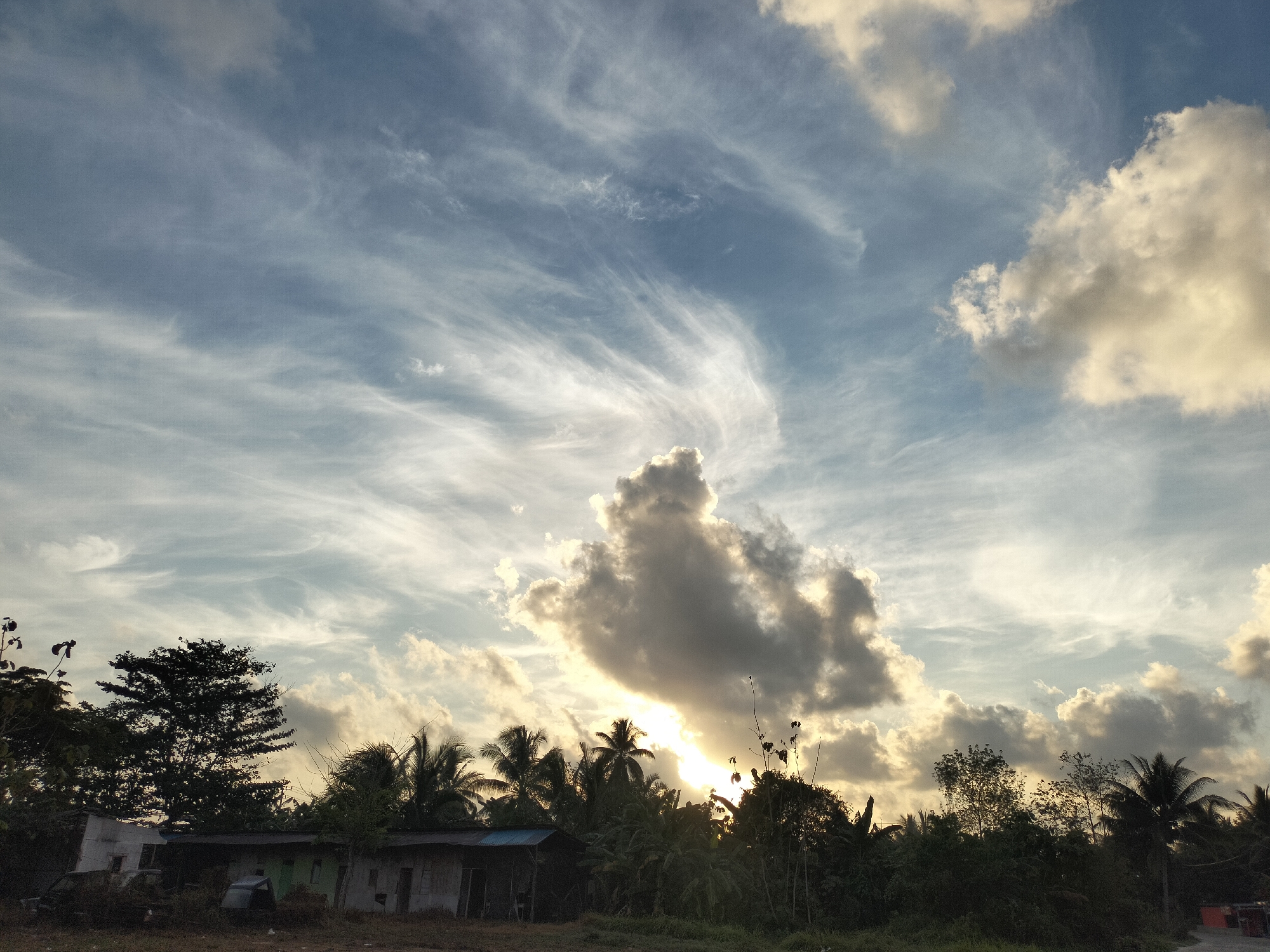 The image depicts a rural landscape at dusk in Karang Indah, Indonesia. The sky is filled with dramatic clouds, with a large, dark cloud group positioned centrally and partially obscuring the setting sun, casting a warm glow. Wispy cirrus clouds streak across the pale blue sky. In the foreground, a line of lush green trees and palm trees forms a dense silhouette against the sky. Below the trees, a low-lying structure, possibly a small house or shed with a corrugated metal roof, is visible. To the left of the structure, an old vehicle is partially obscured by overgrown vegetation. The ground in front of the structures and trees is dry and appears to be rough terrain. The overall impression is one of a tranquil, natural setting at the end of the day.